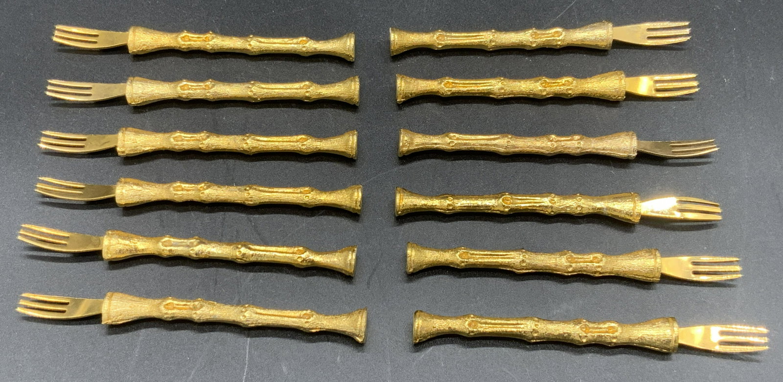 Set 14 Gold Tone Shellfish Forks, Tableware: Fourteen gold toned metal shellfish forks. Each fork has three prongs and a decorative handle. Some undersides possess stickers reading MADE IN HONG KONG. Approx. 3.5L x .5W in. Property of NYC Manhat
