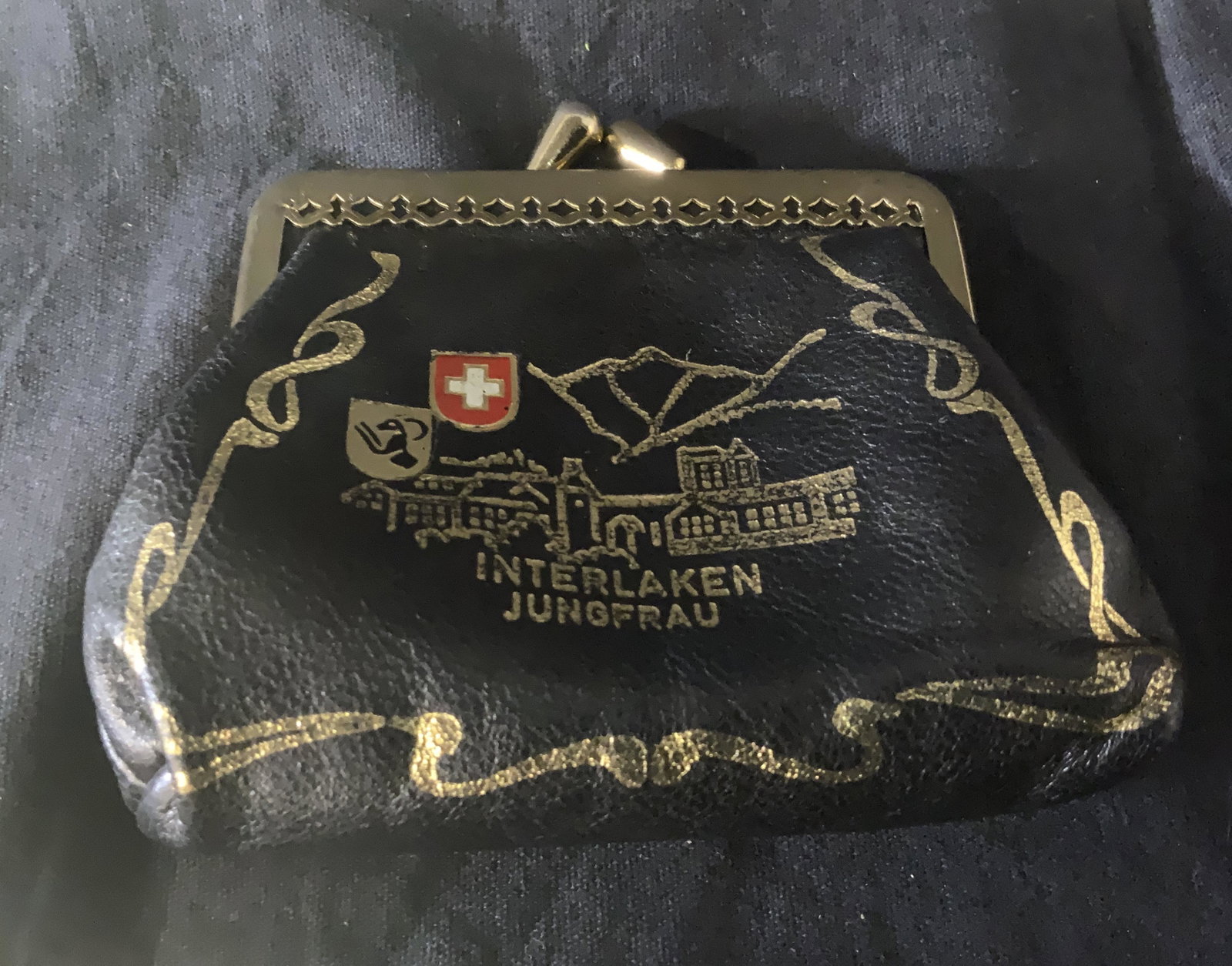 Swiss Army Black Coin Purse: Swiss army black coin purse, exterior of purse displays a townscape the Swiss army symbol and the words ‘Interlaken Jungfrau’, piece measures 4 inches wide by 3.5 inches tall, some wear to piece, 