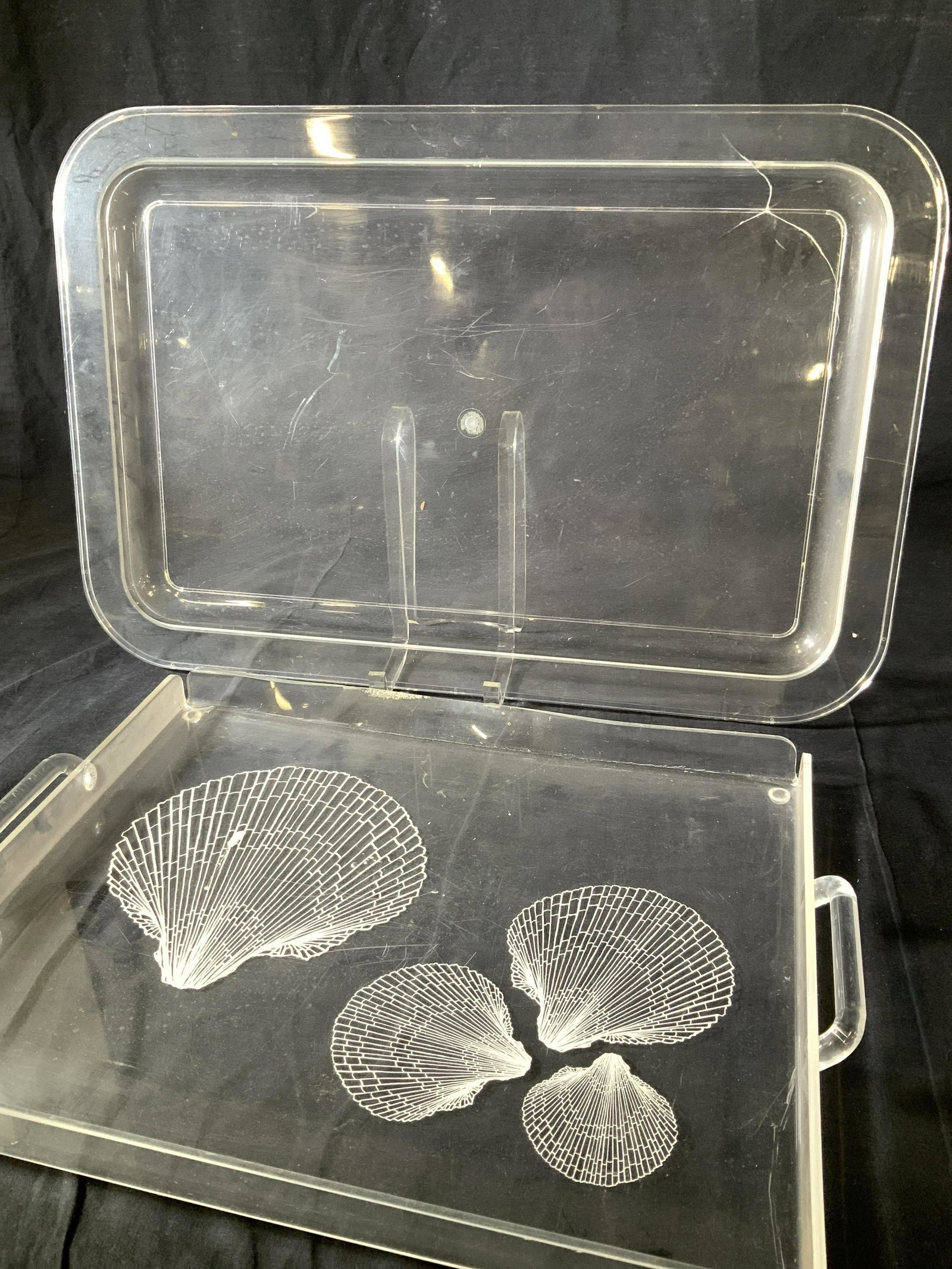 Lot 2 Clear Celluloid Serving Trays: Be achy feel tray with shell decor and carry handles. Side wear ( melted ), 18.5 x 14 in. Other tray measures app 22.75 x 15.25 in. Crack in corner. Works to protect wood table and for casual outdoor 
