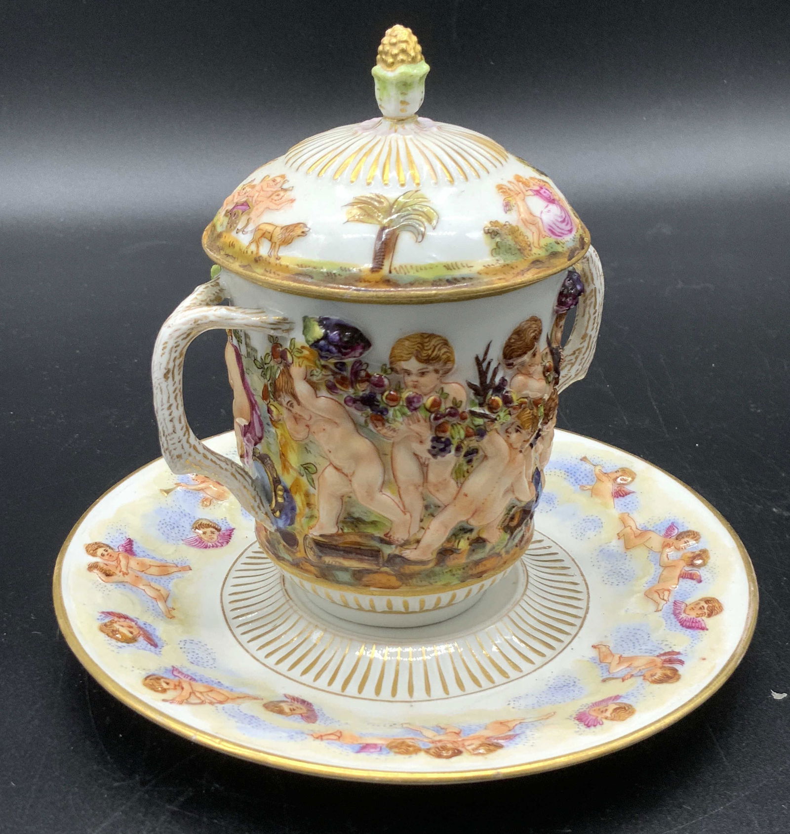 Set2 Antique CAPODIMONTE Porcelain Teacup & Saucer: An antique white porcelain lidded teacup and saucer set featuring hand painted designs. Both pieces depict hand painted cherubs / putti frolicking in a nature scene and amongst the clouds. They are se
