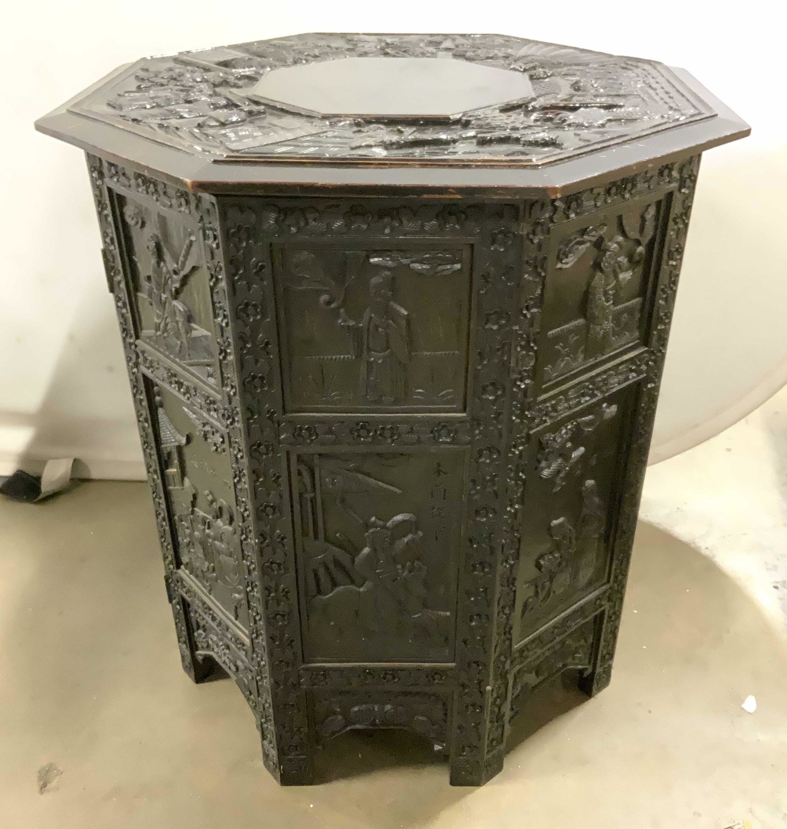 Vintage Asian Carved Wooden Side Table: Hexagonal Asian, wooden side table. Decorated with intricate carving depicting a number of figures. Top is secured to base by latches on its underside. Top can be removed and base can be collapsed for