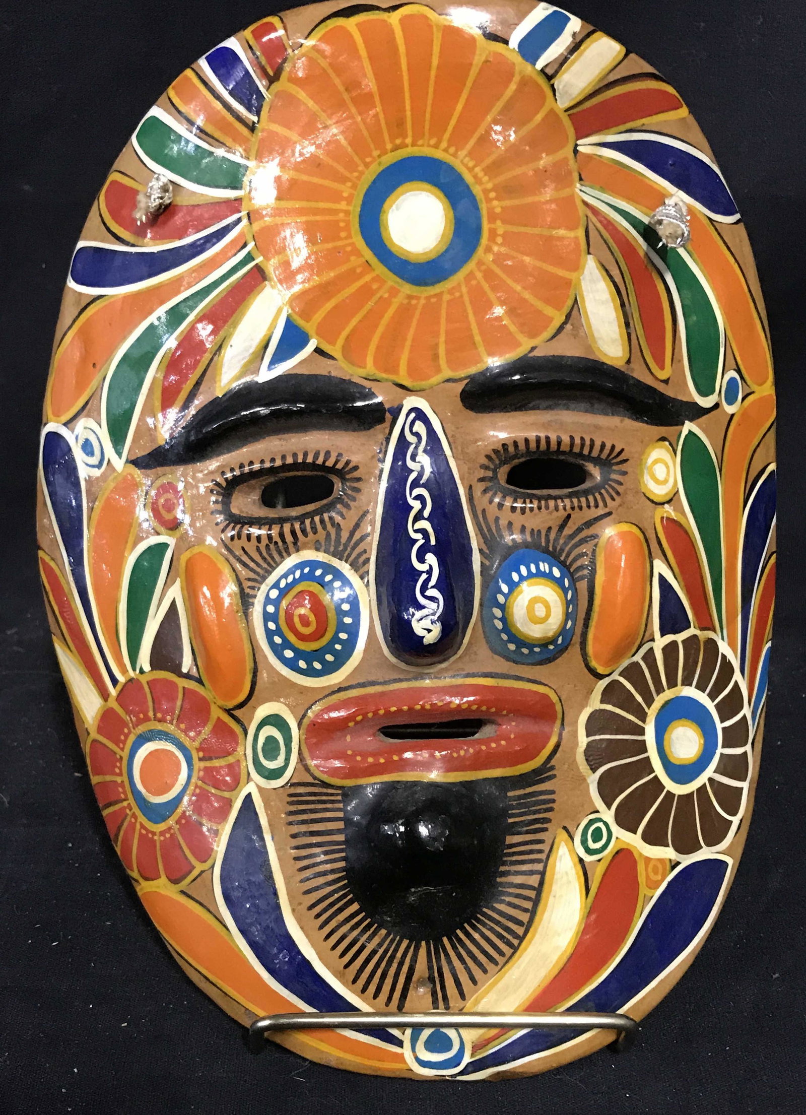 Hand Painted Folk Art Earthenware Mask: Hand painted earthenware decorative mask. Vibrant floral and feather pattern, colored in red, blue, orange, and cream colored tones. A braided string on the reverse of the mask allows for hanging. Mea