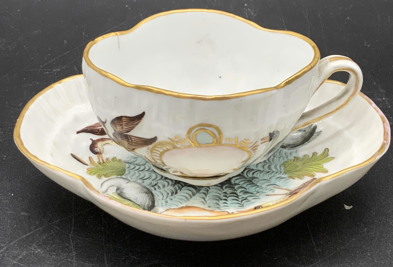 Set 2 Antq CAPODIMONTE Porcelain Teacup & Saucer: A vintage, possibly antique, white porcelain teacup and saucer set featuring hand painted designs. Both pieces depict hand painted birds (herons, swans) frolicking in a nature scene. Gilt line work th