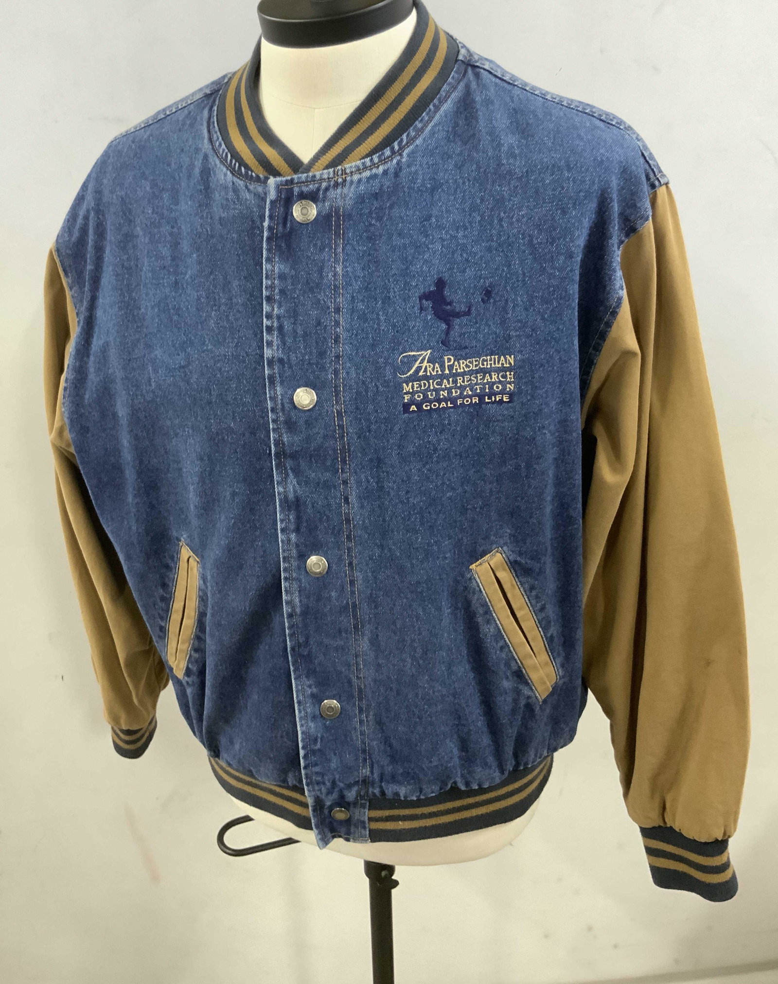 REGIS PHILBIN Dunbrooke Denim Style Jacket: A blue denim style jacket with brown hued cotton sleeves. Closes with silver tone metal snap buttons. Two exterior lined pockets. Piece has a patch on the obverse that reads ARA PARSEGHIAN MEDICAL RES