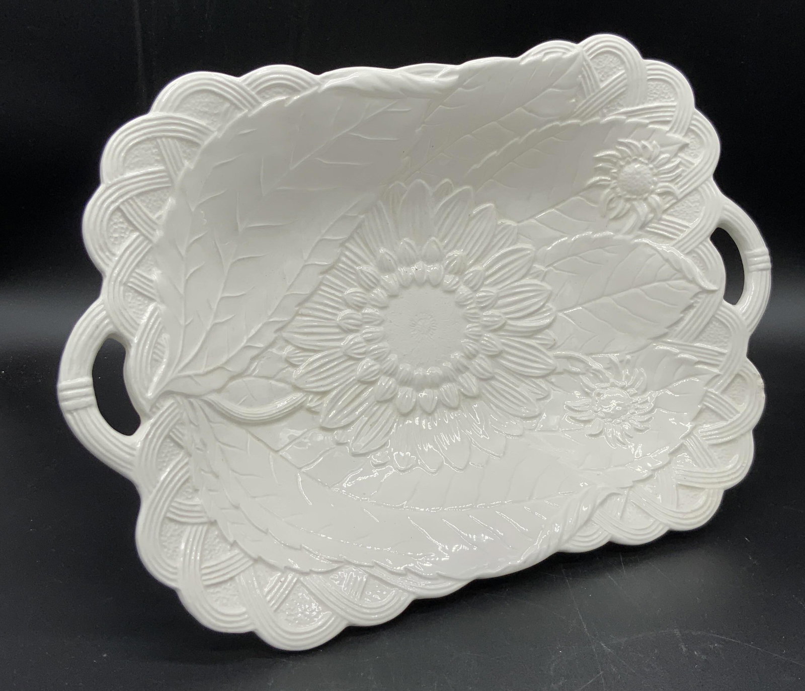 HALDON GROUP White Porcelain Pedestal Dish: A glazed white porcelain pedestal dish with two twin handles on either side. Primarily white in color. Embossed accents throughout depicting florals, sunflowers. Underside is signed THE HALDON GROUP, 