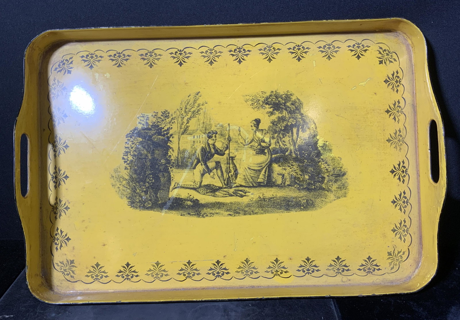 Vintage Yellow Toleware Handled Tray: A vintage painted metal Toleware tray. The piece features twin handles on either side. Primarily yellow with black accents. The center scene depicts a man and a woman in a nature setting. Surface wear