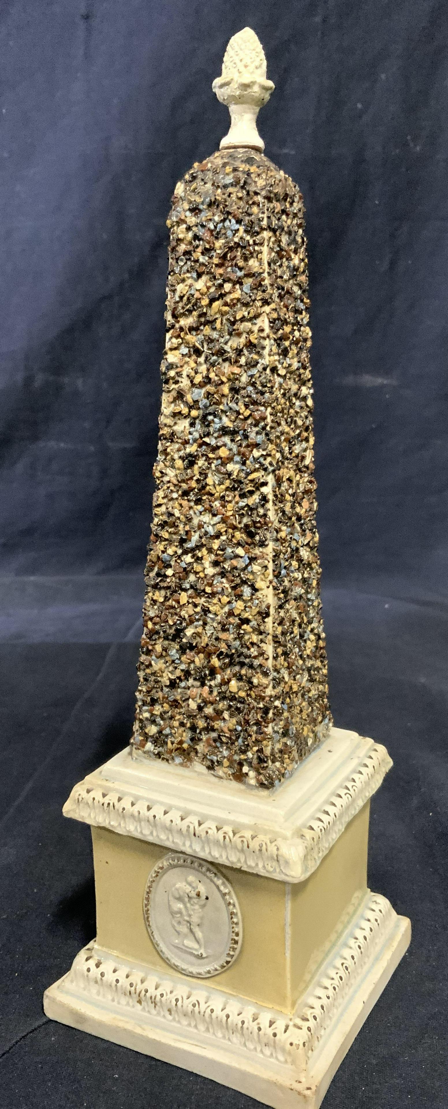 Vintage Ceramic Porcelain Obelisk Ornament: Obelisk appears to be ceramic possibly porcelain. Piece is sand toned . Top of piece has artichoke finial style structure. Near base has oval shaped medallion style structures. Piece measures approx 1