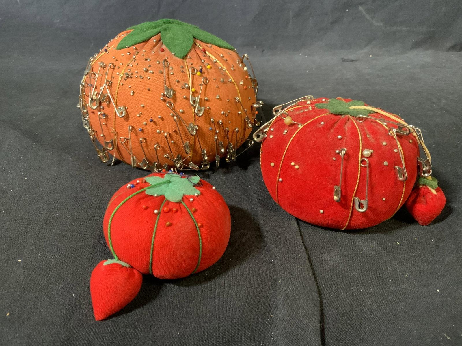 Group Lot 3 Tomato Pin Cushions: Group lot 3 tomato pin cushions, each is a different size with the largest measuring 5 inches tall and 4.5 inches wide, each is covered in sewing pins and safety pins, all have some wear to them possi