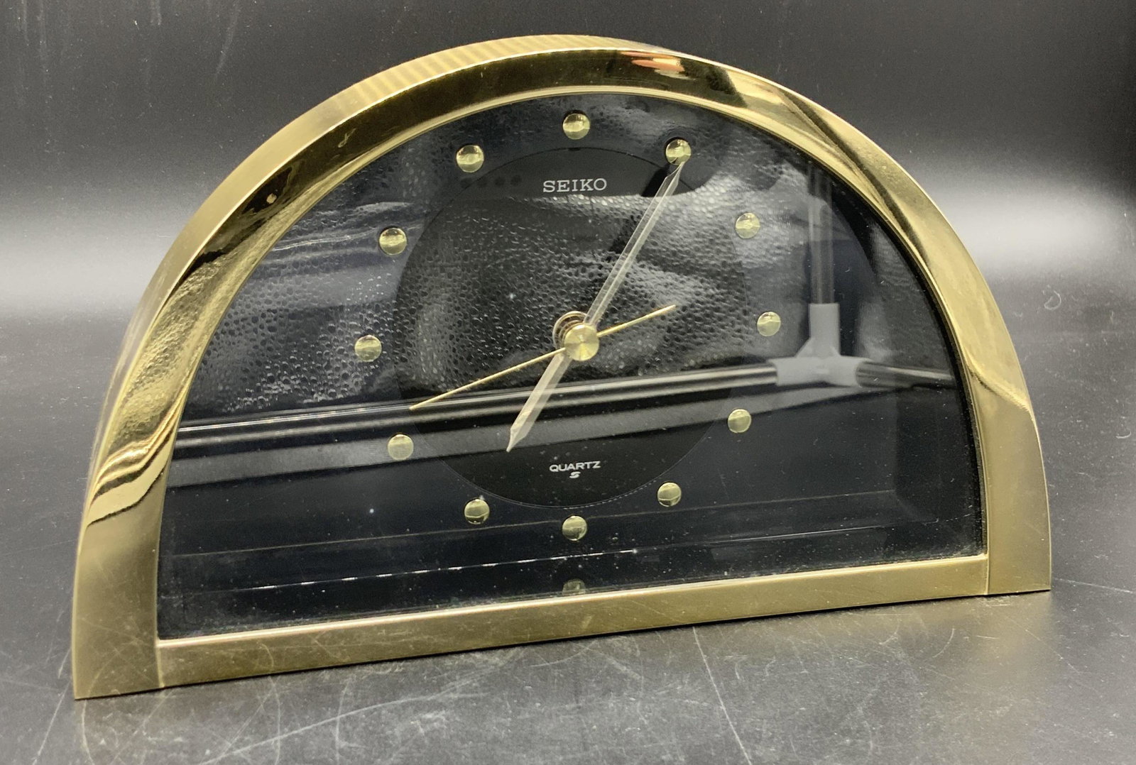 SEIKO Signed Tabletop Quartz Clock: A polished gold tone metal clock with a black clock face and gold & silver toned hands. The face reads SEIKO, QUARTZ. The piece has an arched, semi circle form. Underside has felt. Underside reads REF