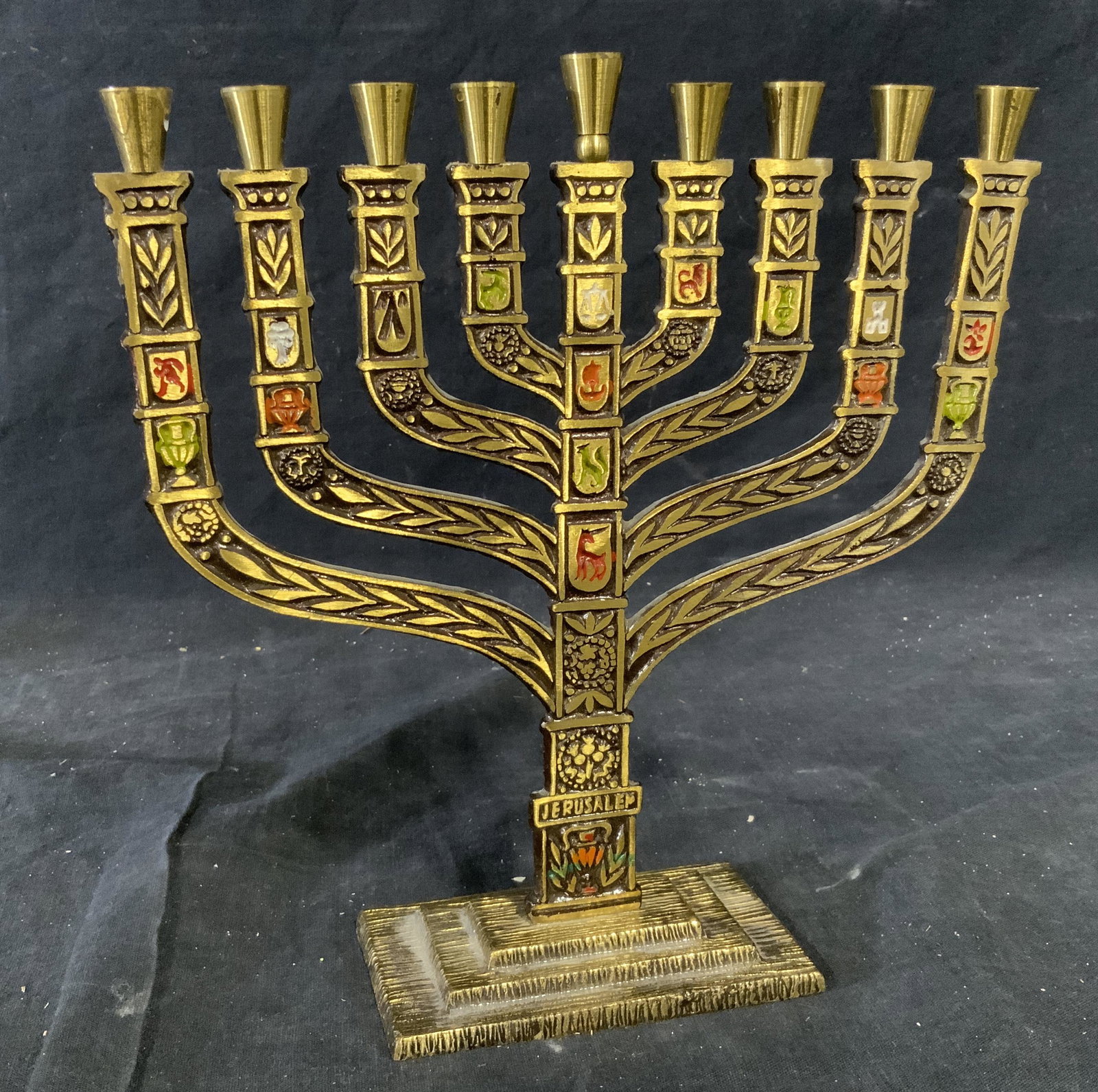 Gold Toned Metal Menorah: Gold toned metal menorah, text to base reads ‘Jerusalem’, subject a gold toned metal menorah with hand painted depictions of animals and symbols, piece measures 8.5 inches tall and 8 inches wide, 