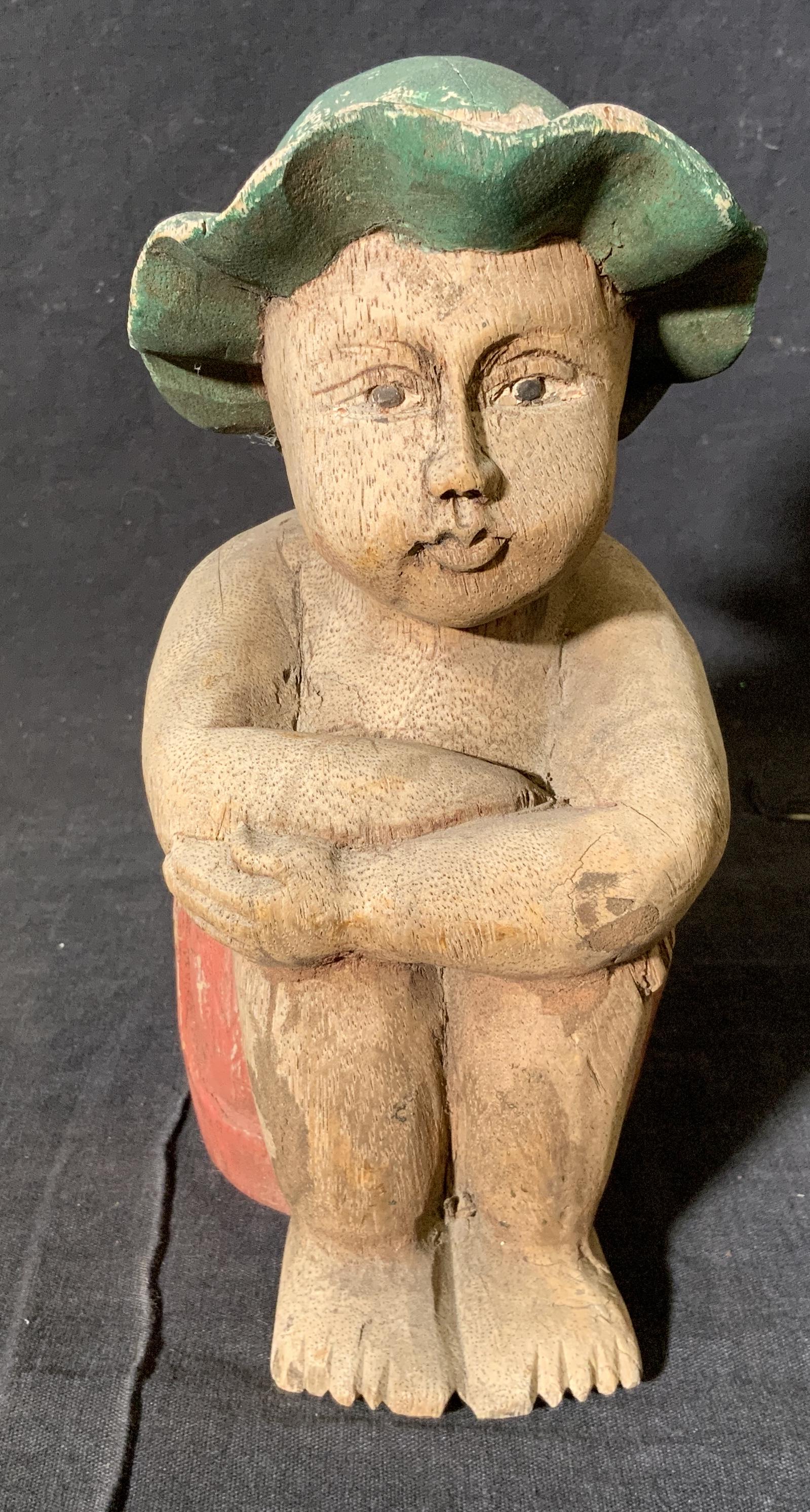 Hand Carved Wooden Figural Sculpture: Hand carved wooden figural sculpture, no apparent inscriptions or markings, subject a young boy in red shorts and green sun hat seated with knees tucked in, figural measures 8 inches at tallest point,