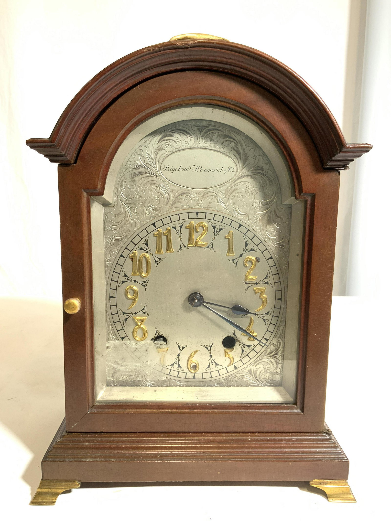 BIGELOW BERNARD CO Vintage Mantel Clock: Clock face reads BIGELOW BERNARD CO. Mantel clock is wooden. Piece appears to be vintage. Piece has windowed door & brass handle at top. Feet are gold toned & brass. Clock face is silver toned with go