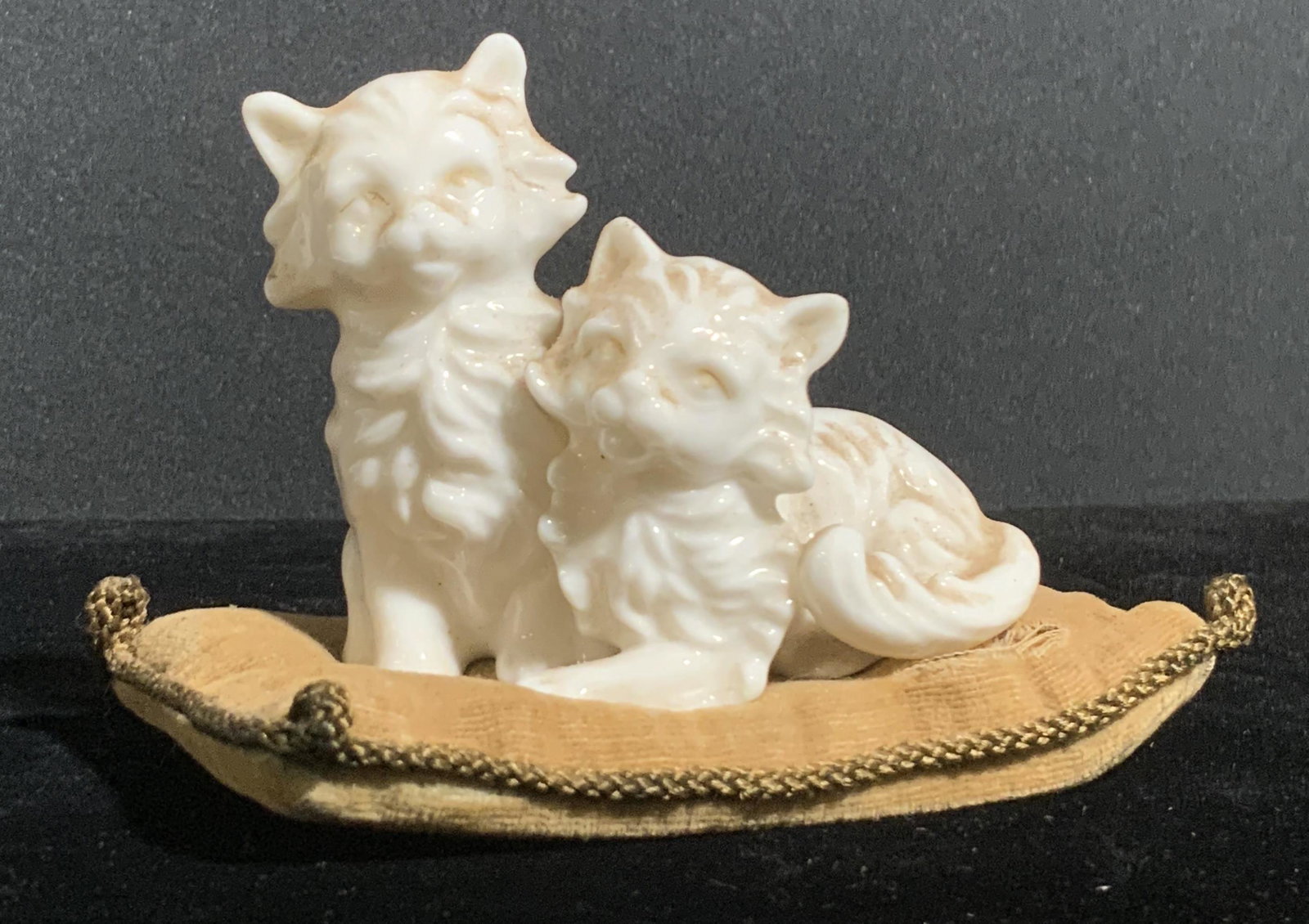 Vintage Porcelain Cat Figural W Fabric Pillow: A white porcelain figural depicting two cats sitting side by side on a pillow. The cats feature embossed porcelain accents throughout. The pillow the cats are affixed to is a light brown hue. Undersid