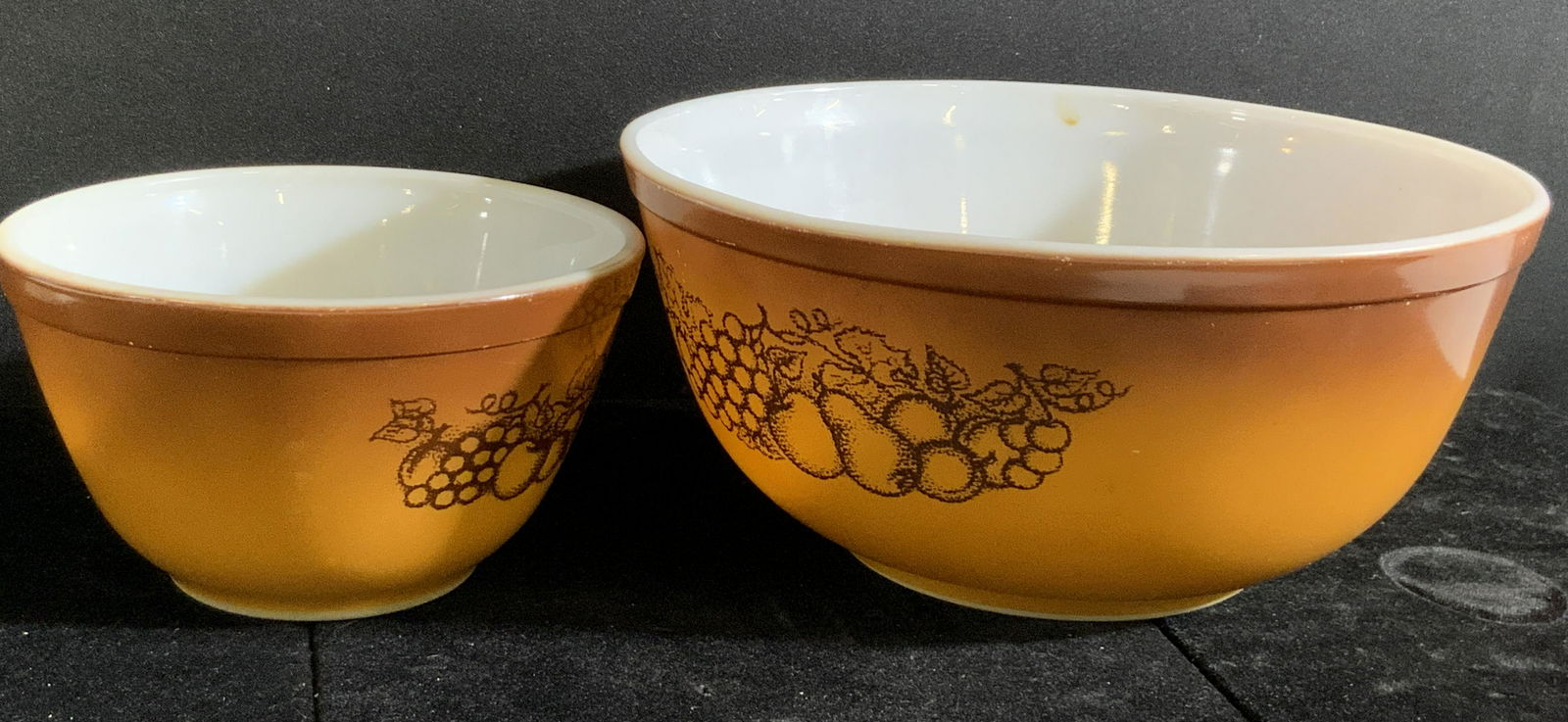 2 Vntg Trademark PYREX Orchard Fruit Pattern Bowls: A pair of vintage Pyrex bowls. Both are a warm ombré brown hue. Both possesses the orchard fruit pattern. Pattern depicts pears, grapes, apples, and more. One bowl is larger than the other. Underside