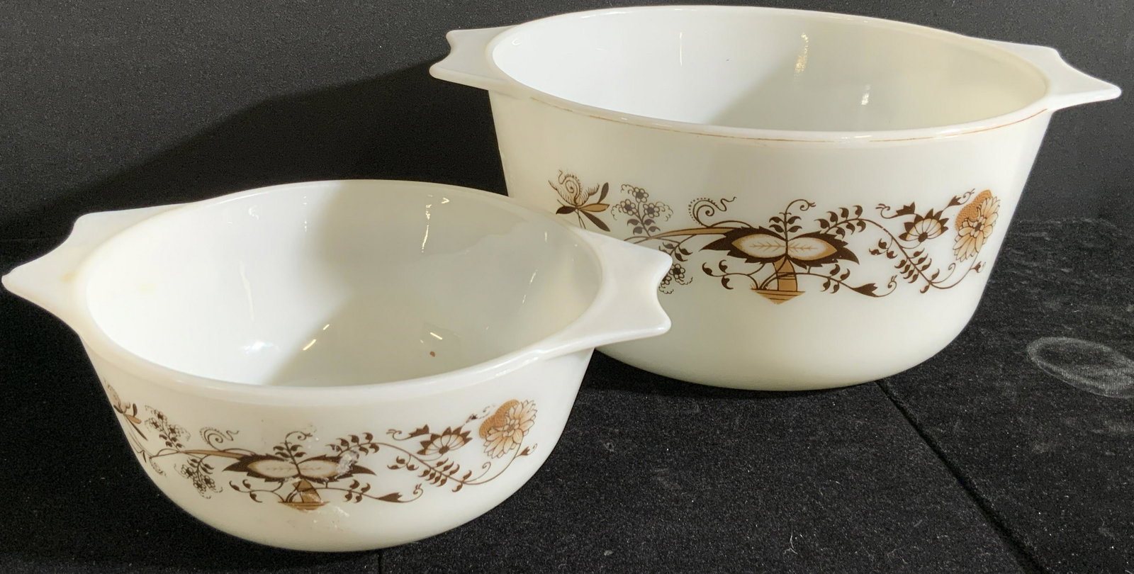 Pair Vntg English PYREX Brown Floral Pattern Bowls: Two vintage white hued Pyrex white hued bowls. Both possess the same pattern and have handles on either side. Undersides read PYREX ENGLAND. Larger bowl approx. 4.5H x 10W in. Both pieces feature some