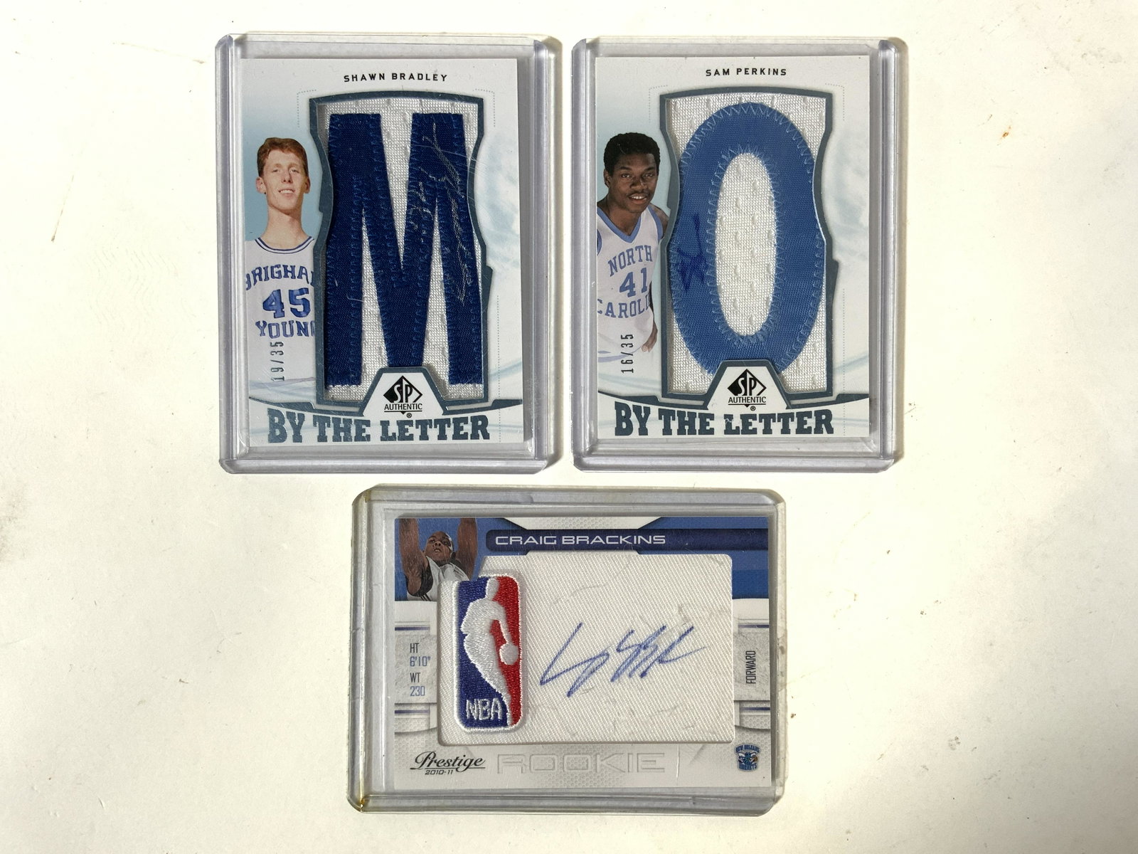 Lot 3 College & NBA Basketball Cards: Lot includes 3 cards including Upper Deck By The Letter Shawn Bradley, Upper Deck By The Letter Sam Perkins & Craig Brackins Panini no. 21. Each card is in plastic casing. Measures approx 3.5 inches t