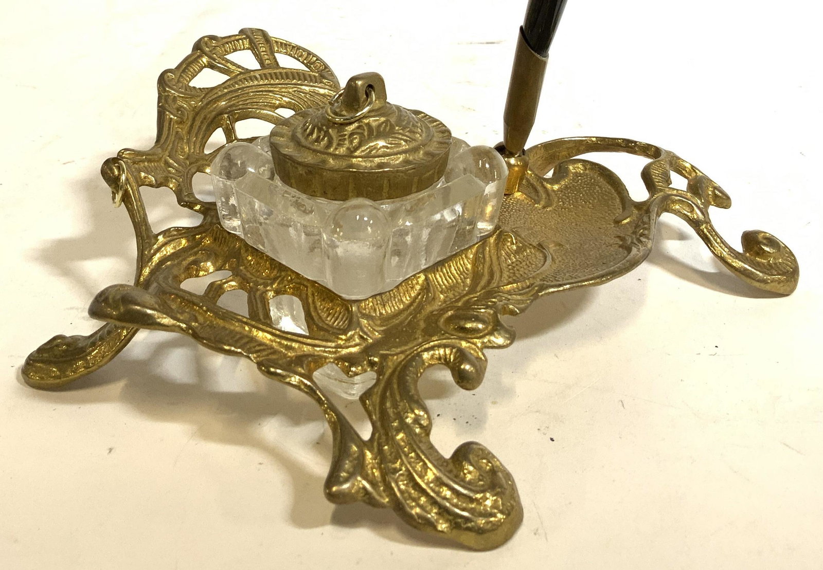 Vintage Brass Inkwell W Pen: Inkwell is vintage and brass. Piece is footed. Contains ornately detailed structures. Inkwell has glass basin with brass lid. Lot includes black toned pen. Piece With pen in holder measures approx 7.5