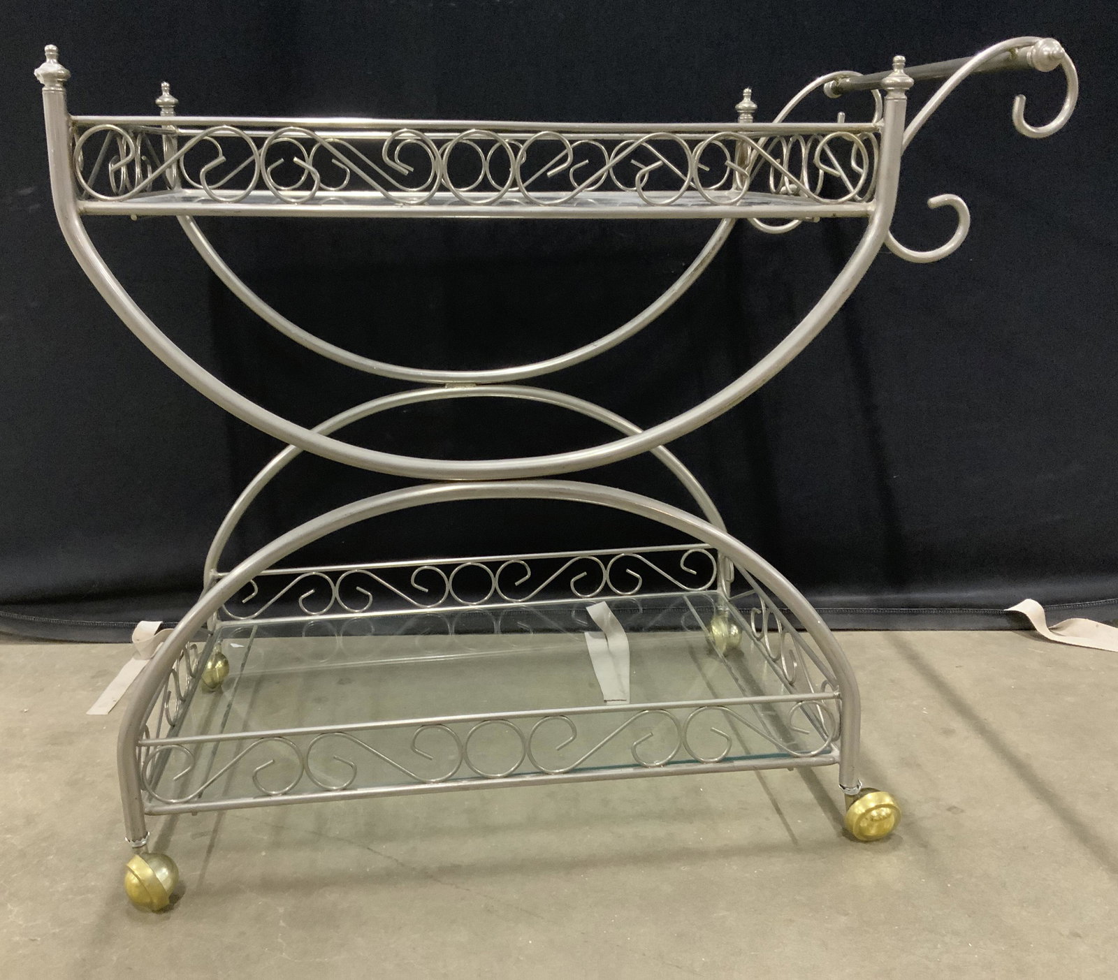 Vintage Scrolled Metal Bar Cart w Casters: Vintage bar cart on castors. Bar cart frame is metal, scrolled detailing decorate upper and lower tiers. Castors appear to be brass. Upper and lower tiers house glass panes, act as shelves and trays. 