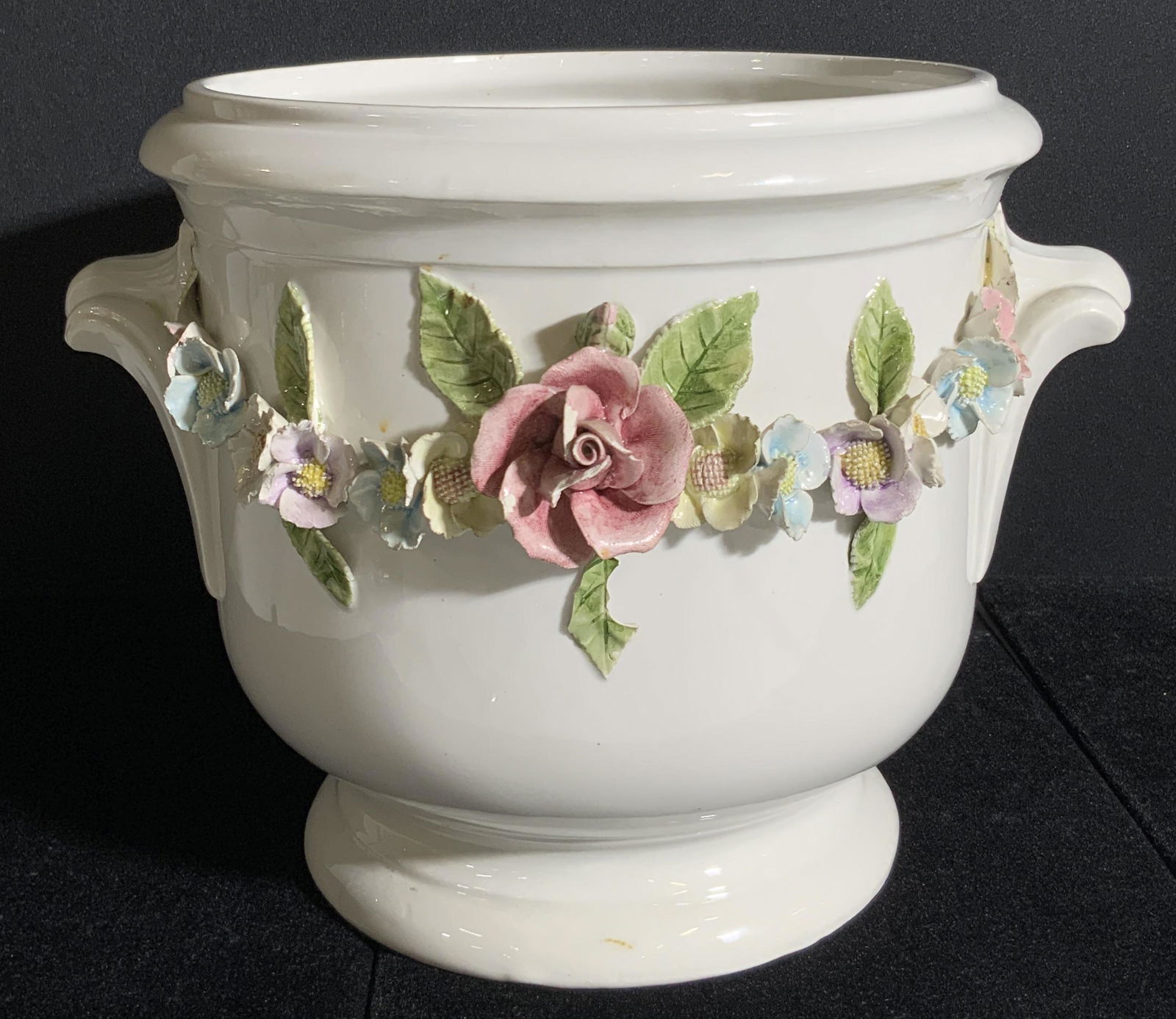 RUBEL Signed Portuguese Porcelain Floral Cachepot: A white porcelain planter with handcrafted raised floral accents. Florals are hand painted and depict roses amongst leaves and other flowers. Primarily blue, purple, red, and green tones accent the fl