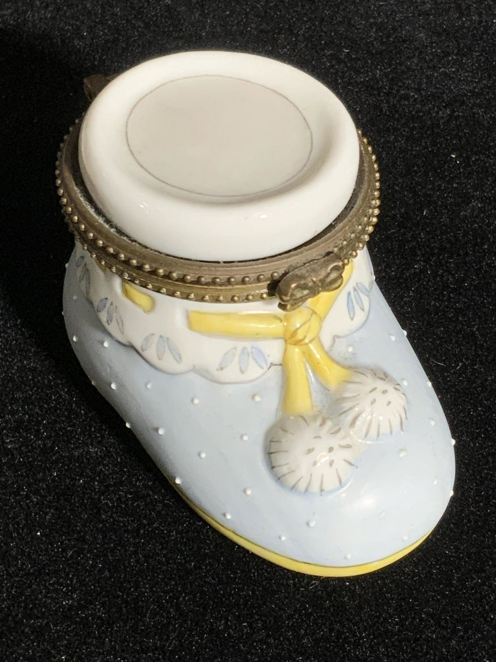 Baby Blue Infant Boot Form Porcelain Trinket Box: A porcelain trinket box in the form of a baby / infant boot. Baby blue hued with white and yellow accents. Raised porcelain details. Piece opens with antiques gold hued hinge hardware. Approx. 2H x 3W