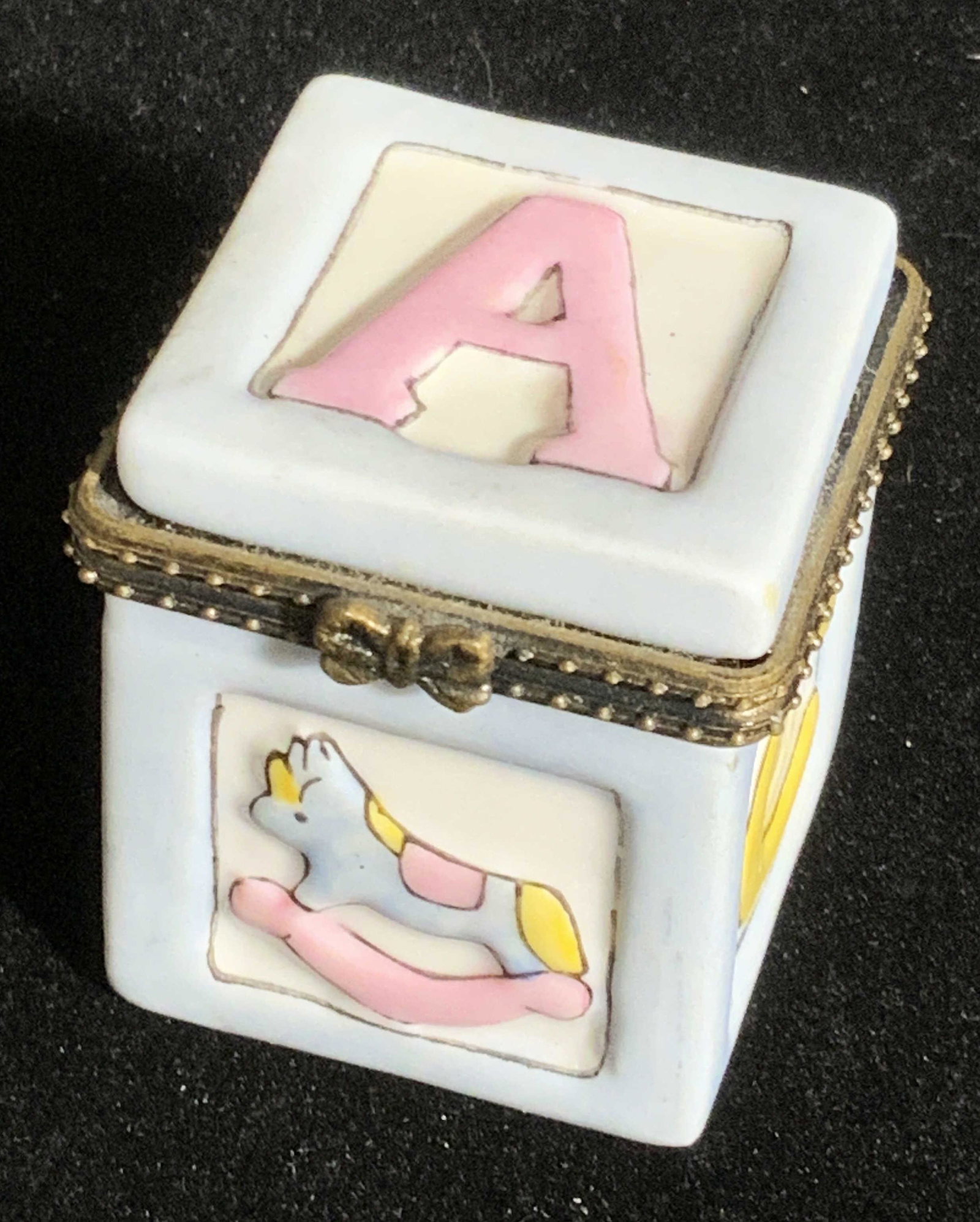 Toy Block Form Porcelain Trinket Box: A porcelain trinket box in the crafted into the form of a toy block. Baby blue hued with white, pink. and yellow accents. Raised porcelain details. Piece opens with antiques gold hued hinge hardware. 
