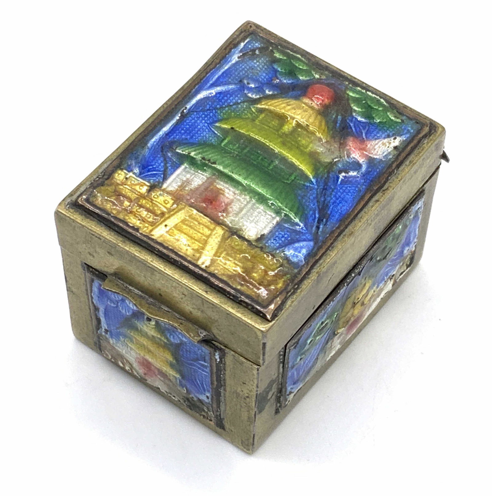 Collectible Enameled Metal Trinket Box: An enameled metal trinket box depicting images of buildings in nature scenes along the obverse and sides. The buildings are raised. The piece is an antiqued gold hue. Enameled scenes are green, blue, 