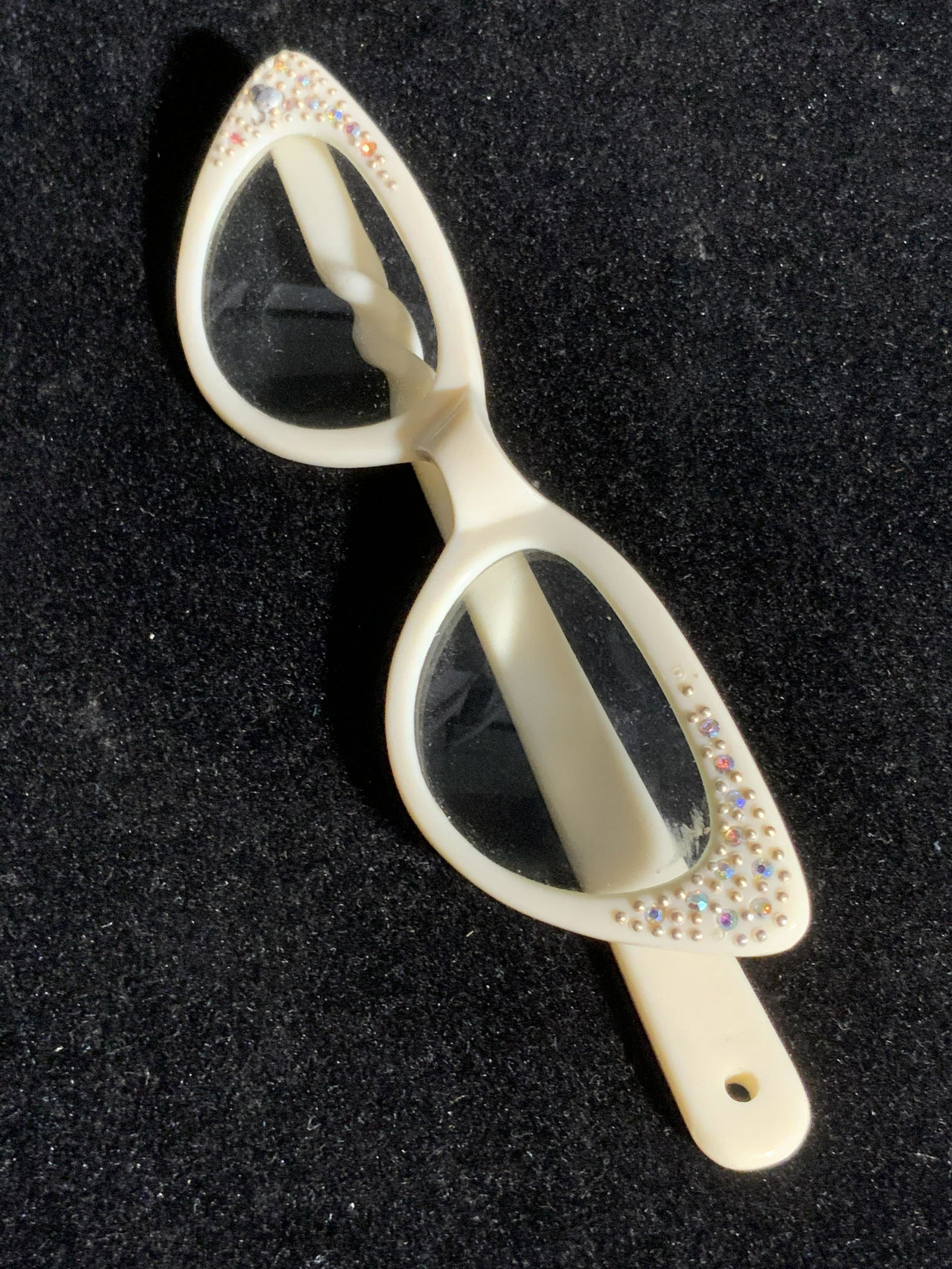 Retro White Cat Eye Rhinestone Glasses, Eyewear: A pair of white hued cat eyes style glasses. The edges are bejeweled with rhinestones and raised accents. One rhinestone missing. Lenses appear to be prescription. Pieces have a handle connected to on