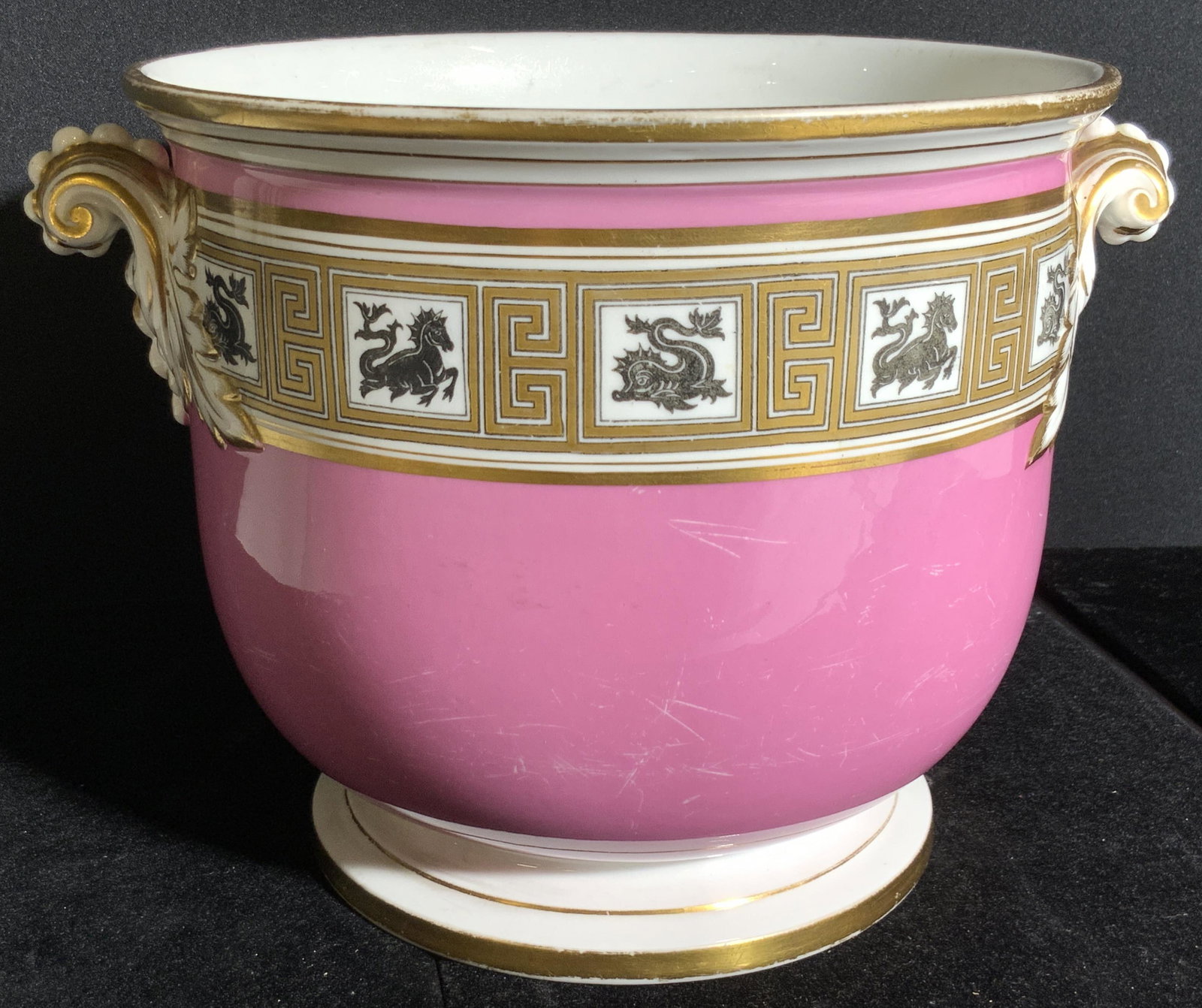 Pink Porcelain Patterned Cachepot: A white porcelain cachepot painted with gilt accents and pink tones. The piece has twin handles with embossed porcelain accents and gilt hues. The patterns feature black mythological creatures that re