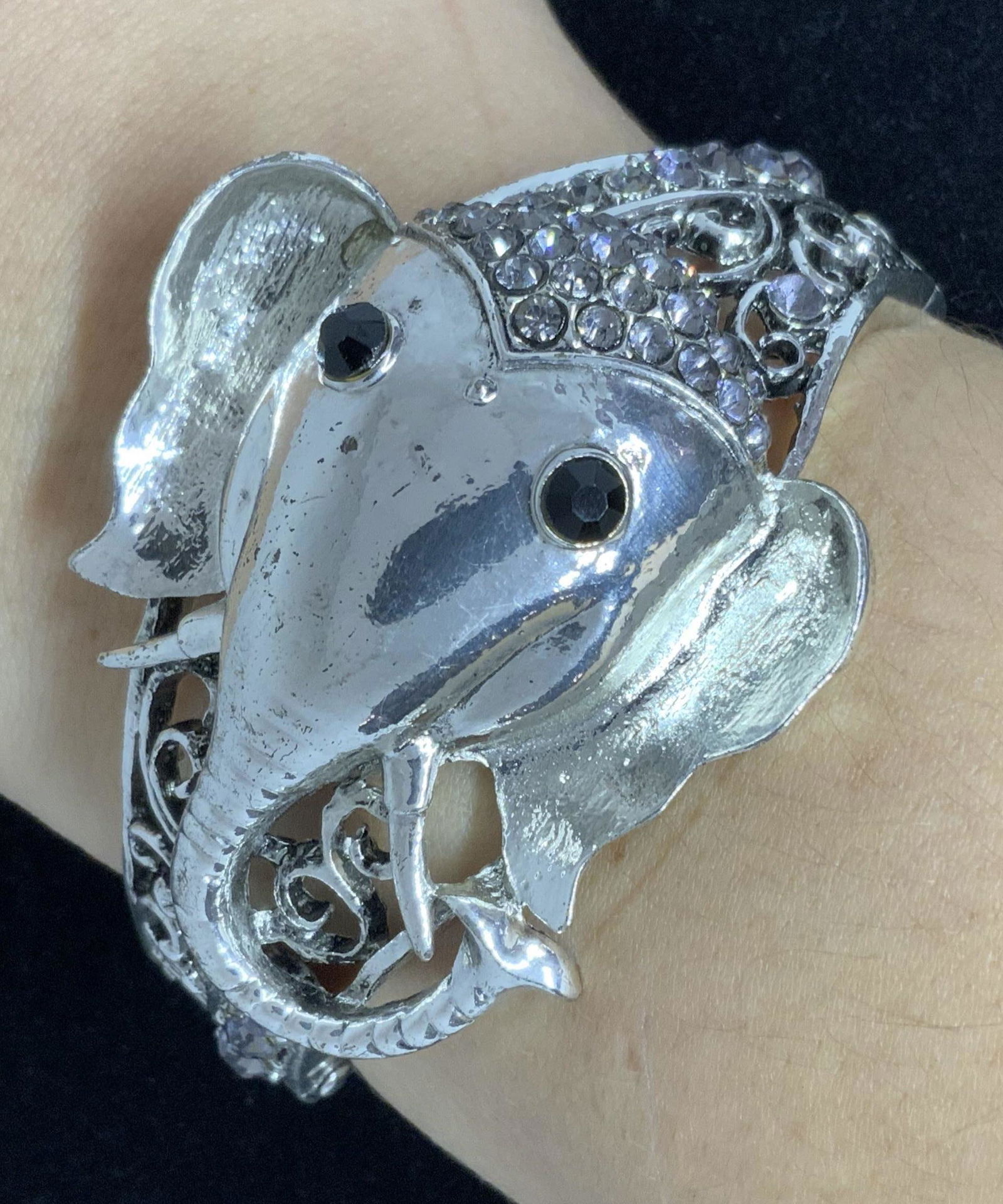 Elephant Form Pierced Metal Crystal Hinge Bracelet: A pierced and etched silver toned metal hinge bracelet. The form of an elephant’s head rests on an intricately pierced band. The elephant has two black, faceted crystal eyes and colorless, faceted c