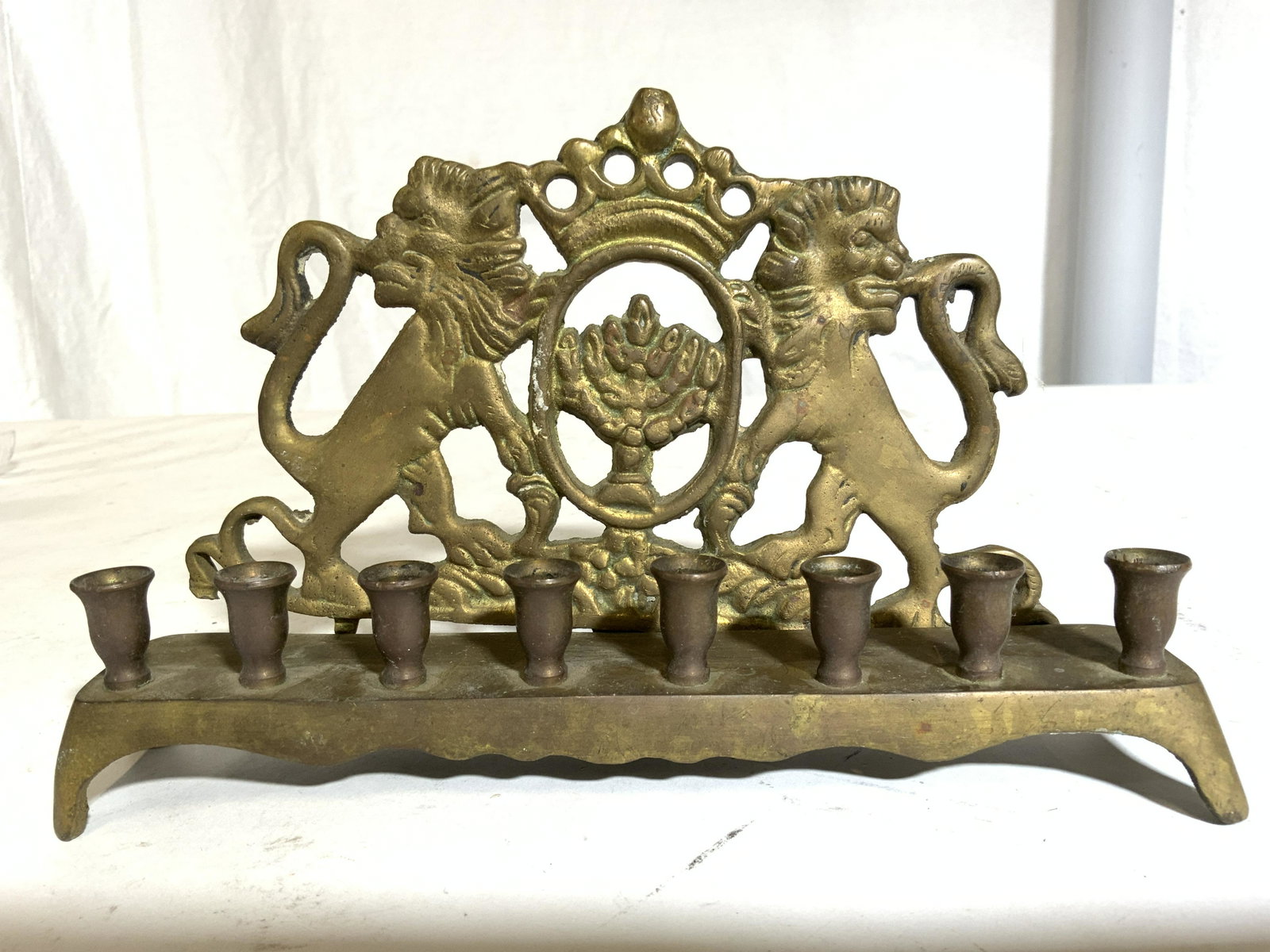 Gold Toned Brass Menorah W Lion Detail: Menorah is vintage & brass. Piece has 8 candle areas. Menorah has mirrored Lion structures on sides along with crown shaped structure with oval structure underneath. Piece measures approx 5 inches tal