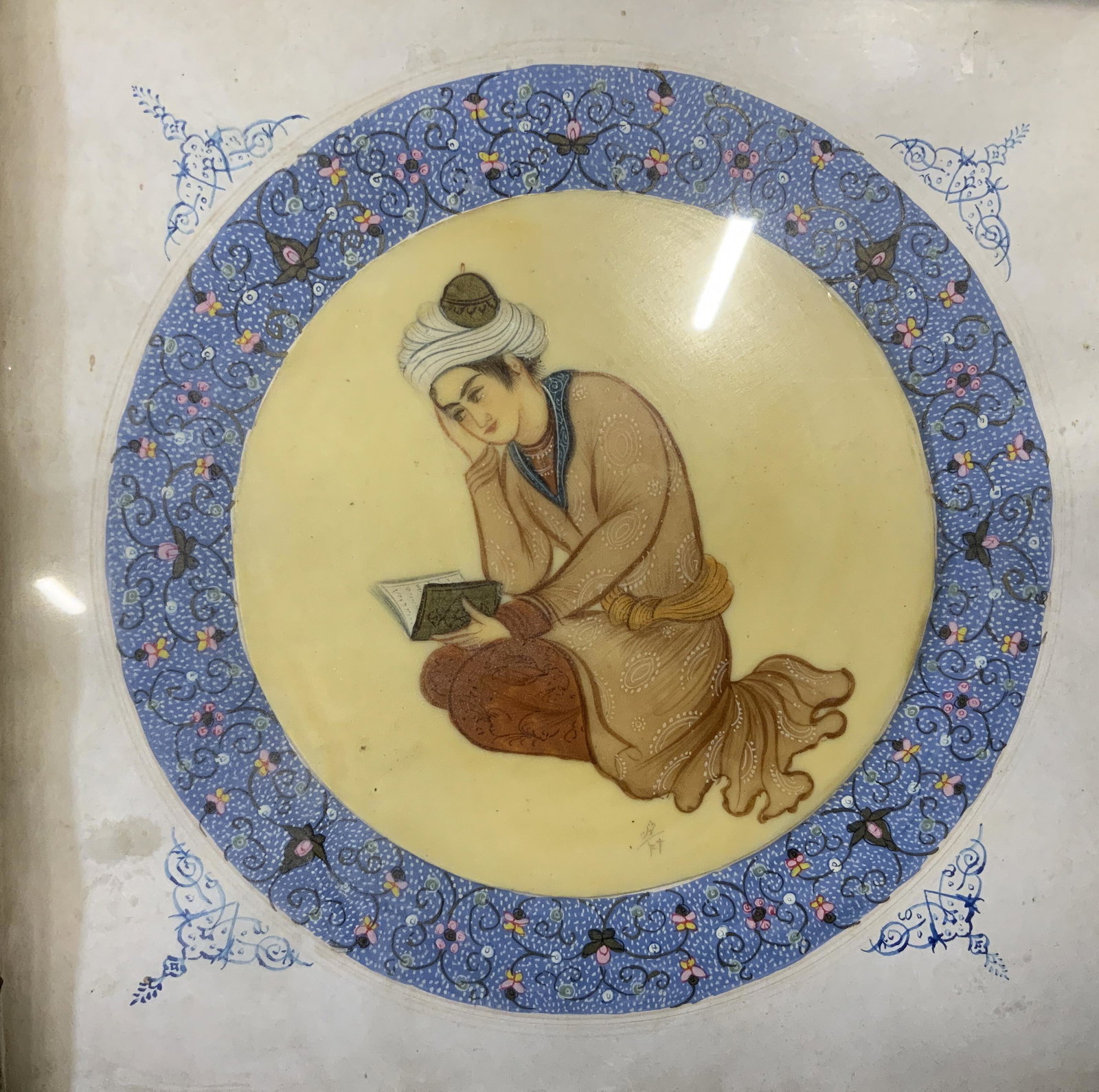 Arabic Painting on Resin: Arabic painting on resin, resin possibly alabaster, Arabic text to the lower right corner, subject a man seated on his knees reading a book, decorative floral matting, wooden frame with geometric patt