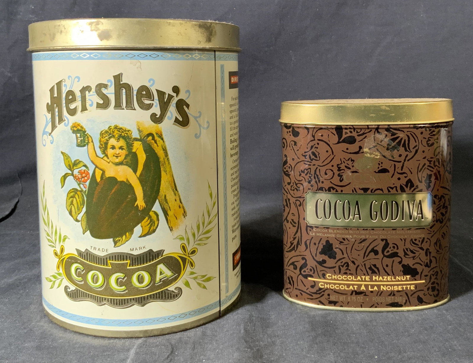 Lot 2 Hershey & Godiva Cocoa Containers: Lot 2 Hershey & Godiva Cocoa Containers, inscriptions read ‘Cocoa Godiva Chocolate Hazelnut’ and ‘Hershey’s Trademark Cocoa’, smaller container measures 5 3/8 inches tall x 4 1/2 inches wide