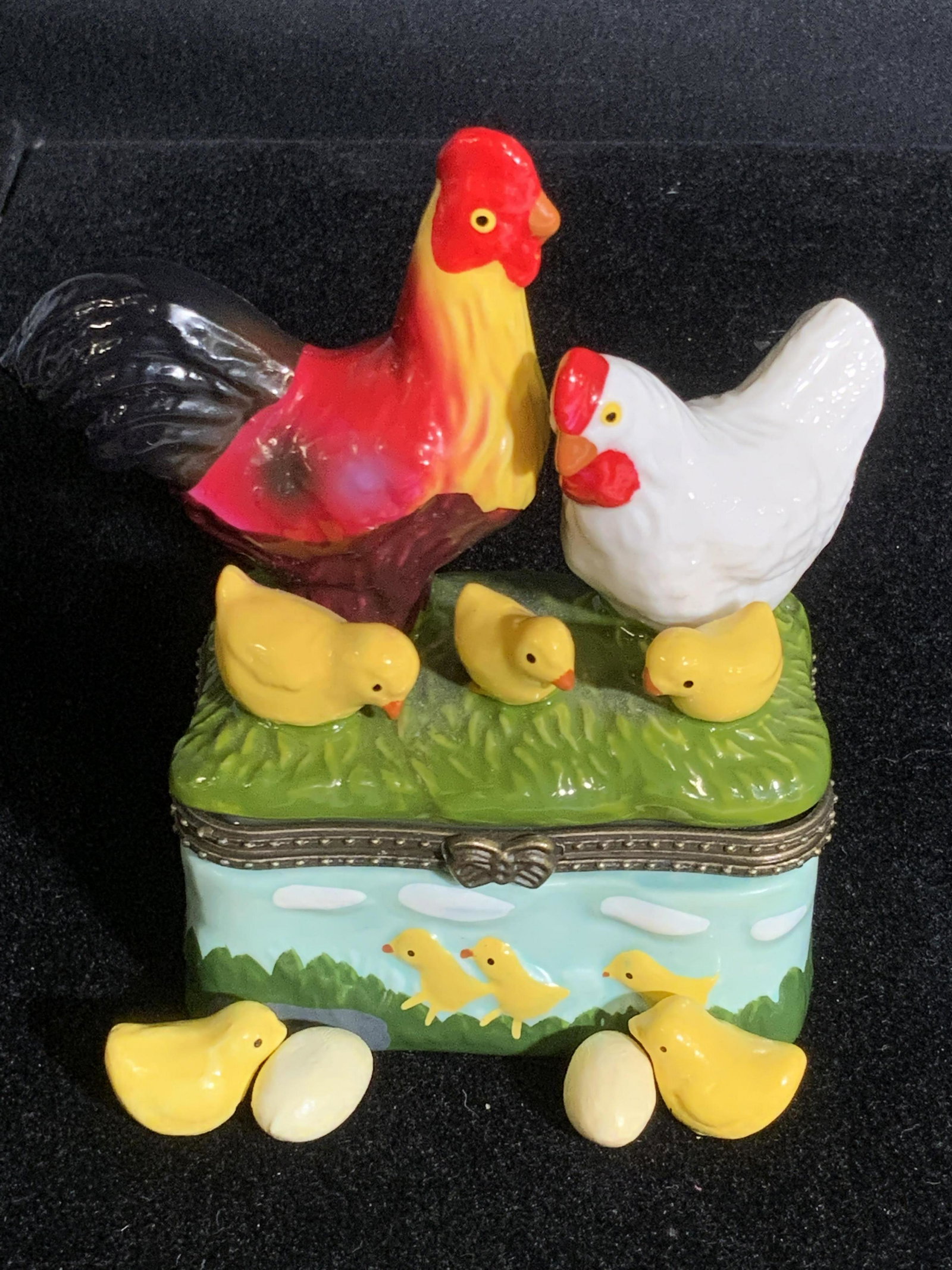 Hand Painted Porcelain Chicken Family Trinket Box: A porcelain trinket box that has been decorated with raised chicken figurals. Upon the lid sit two chicks, a rooster, and a hen. The exterior is a painted nature scene with raised accents. Chicks are 