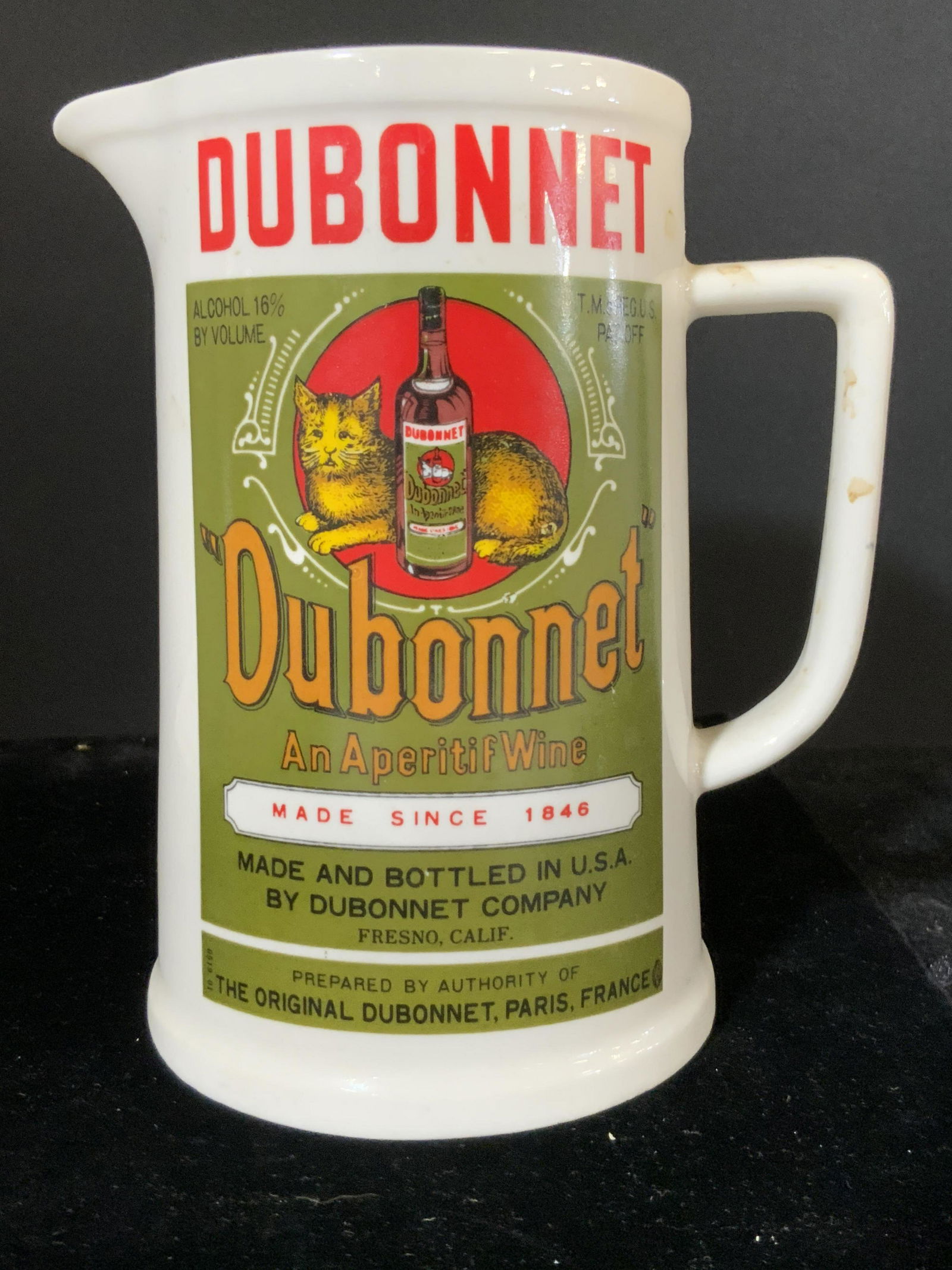 Vintage DUBONNET Cat Motif Porcelain Pitcher: A vintage white porcelain pitcher. The piece reads DUBONNET, AN APERITIF WINE. A cat is depicted laying down next to a bottle of wine. Piece is primarily white, red, and green hues. Approx. 6H x 5.25W