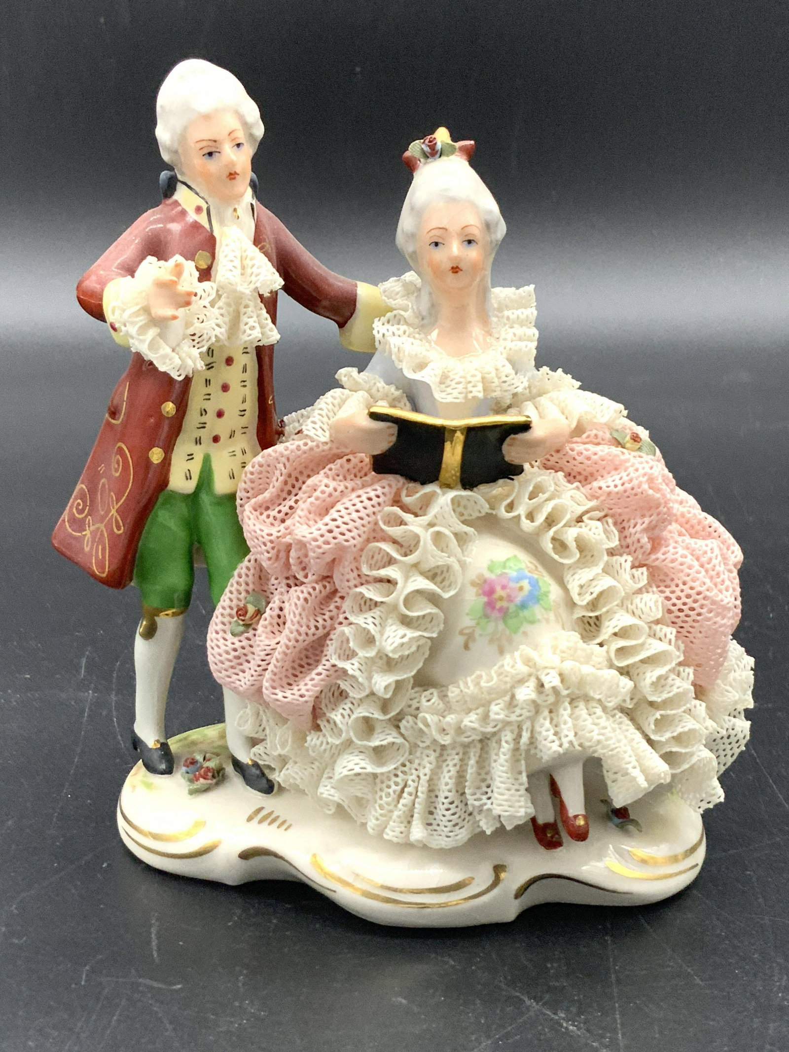 Signed MEISSEN German Porcelain Figural: A figural depicting a man and woman wearing traditional, fine clothing. The male is standing beside the female, who is seated and reading. The porcelain work is hand painted with florals and gilt line