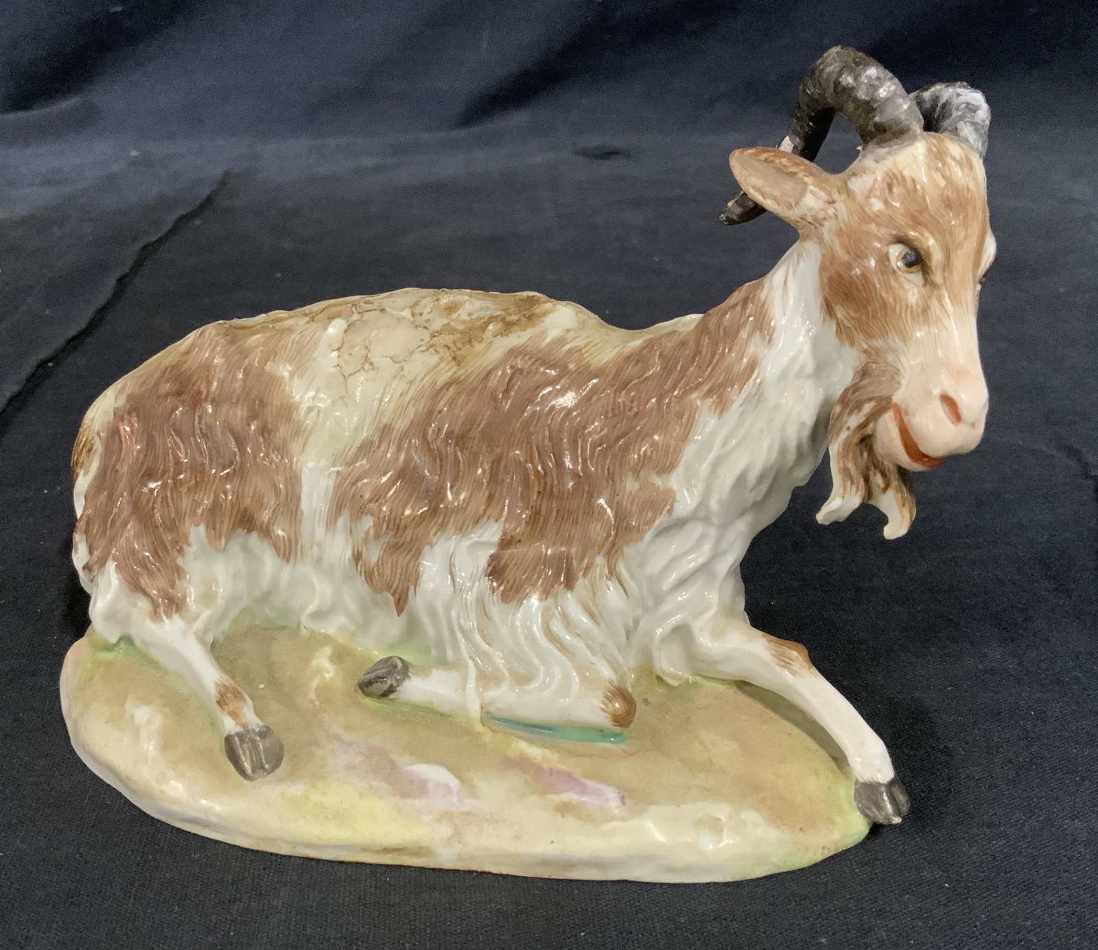 Hand Painted Porcelain Goat Figural: Hand painted porcelain goat figural, label to underside faded from age possibly reads ‘Goat Early Missan’, subject a goat lying down with brown and white fur, surface wear to figural, measures 3.5