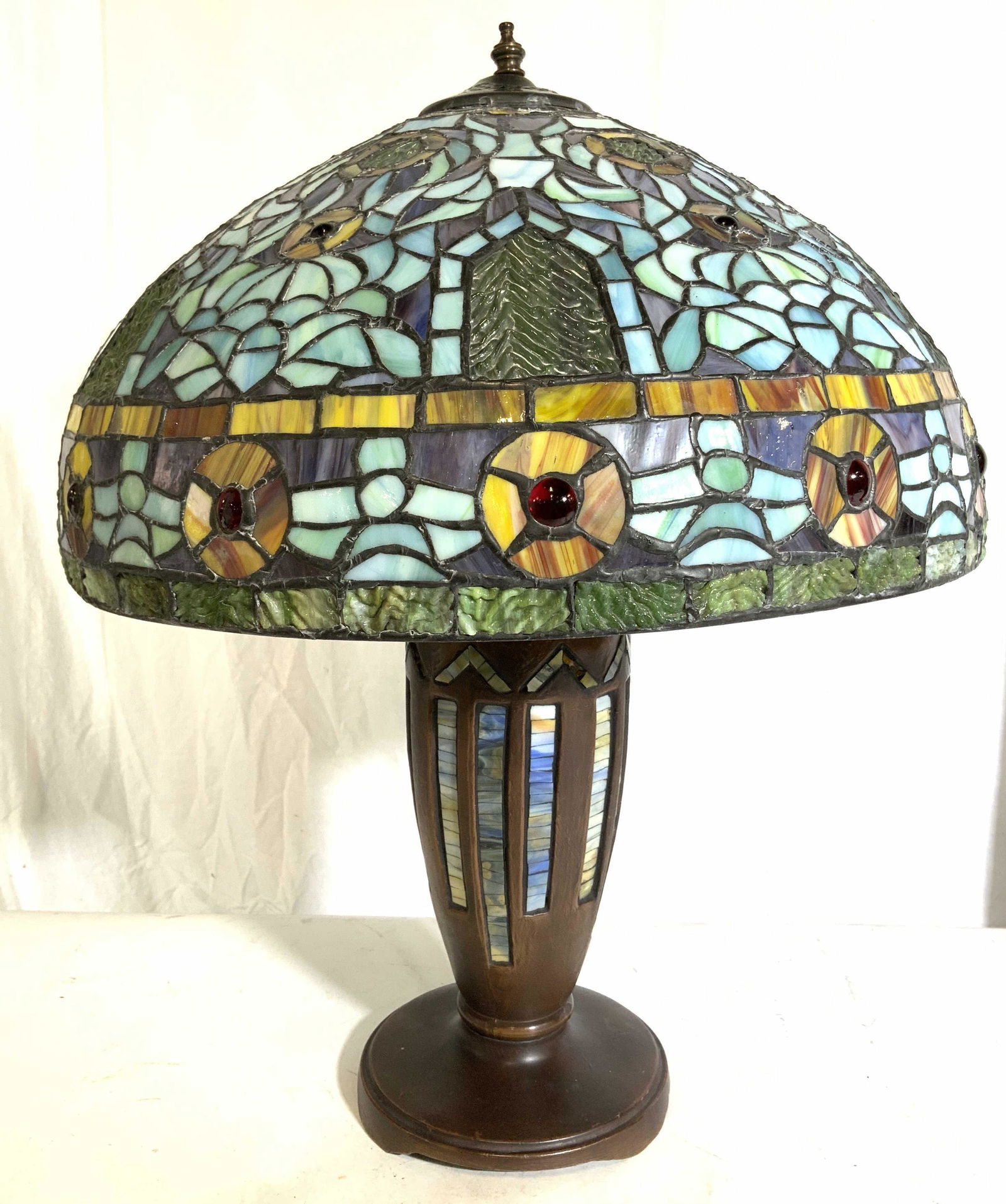 Tiffany Style Stained Glass Lamp W Shade: Lamp is Tiffany styled. Piece has multi toned stained glass, slag glass structures & detail. Lamp base appears to be composite, possibly wooden. Top of piece has bronze toned metal finial. Piece measu