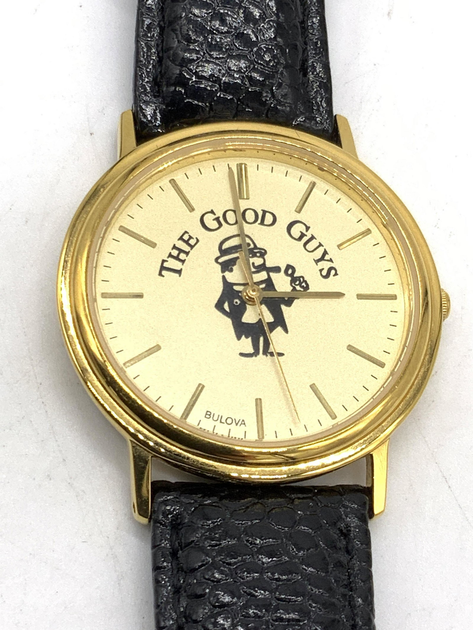 BULOVA The Good Guys Genuine Leather Watch: A black genuine leather wristwatch crafted by BULOVA. Watch face comprised of gold tone metal. Image in the center depicts a man smoking a pipe. Reads THE GOOD GUYS, BULOVA. Watch face approx. 1 x 1 i