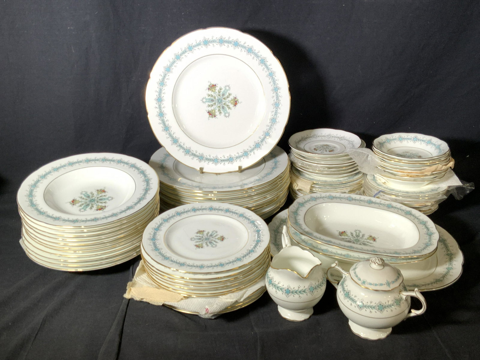 Set 69 COALPORT ‘Geneva’ Bone China Dinnerware: All pieces possess the same fruit basket and blue floral motif pattern with gilt rims. Some pieces have a more muted coloration. Pieces possess some wear consistent with use, discoloration and wear to