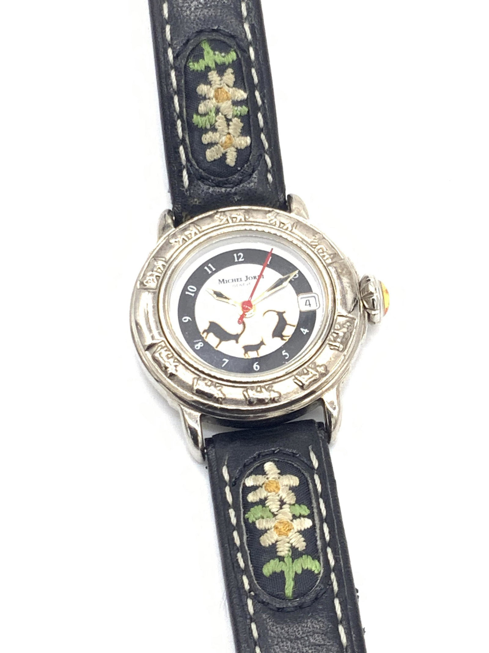 MICHEL JORDI GENEVE Ladies Floral Wristwatch: A Michel Jordi Geneve ladies wristwatch. Crafted from a black leather style material and accented with stitched florals. Florals resemble daisies. Watch face is decorated with long horned goat silhoue