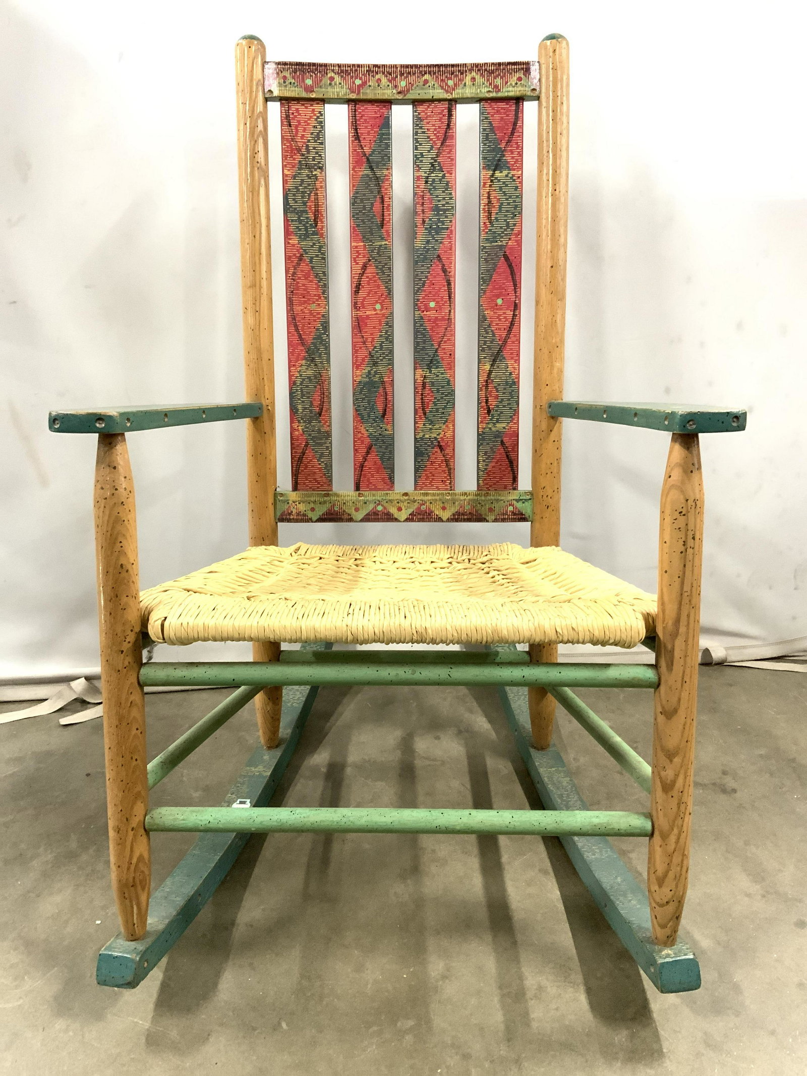 Vintage Wooden Rocking Chair W Rattan Seat: Rocking chair is vintage and wooden. Piece has woven rattan seat. Piece is painted with red and green toned detail. Piece measures approx 39 inches tall 25 inches wide 30 inches deep. Rocking chair, w
