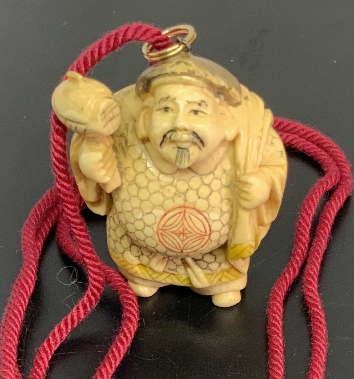 Vintage Signed Asian Carved Bone Netsuke Necklace: A vintage carved bone, possibly resin, Netsuke depicting a bearded man holding a hammer and a sack. The piece is adorned with a bail and affixed to a red cord. Red cord is tipped with metal accents ma