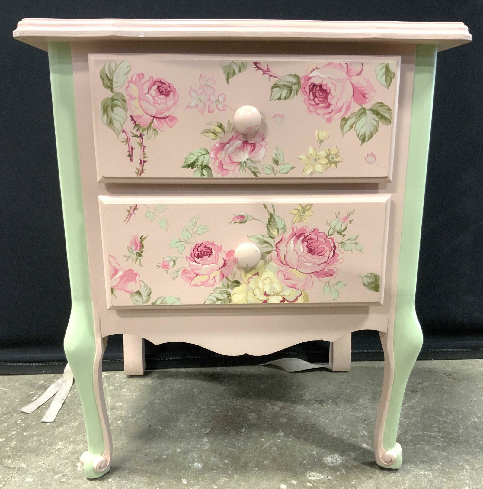 Floral Detailed 2 Drawer Wooden Nightstand: Nightstand has pink and green toned floral, vine, and leaf detail. Piece has 2 drawers each with knob style handles. Front 2 feet have curled detail. Top f piece have floral and leaf detailed pattern.