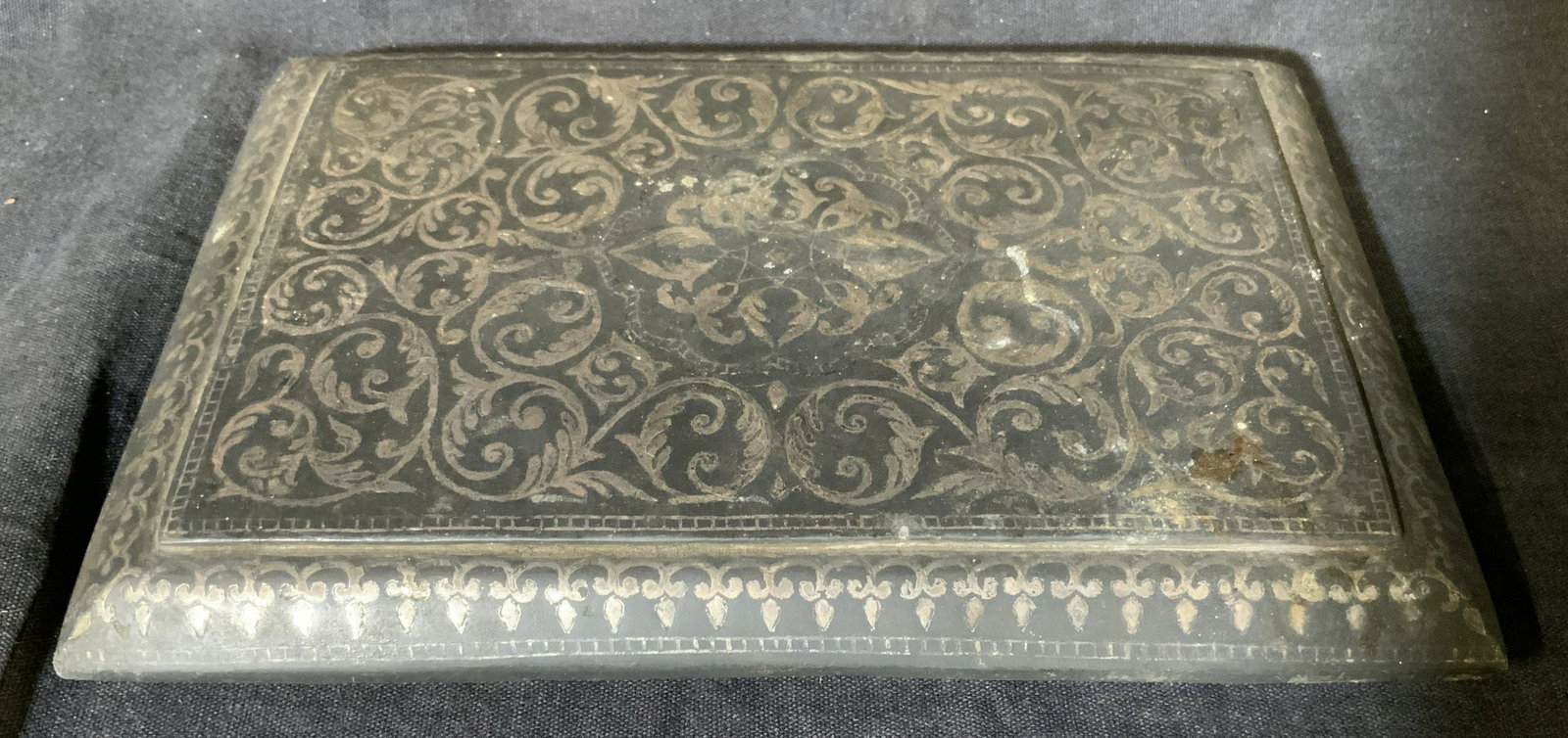 Antique Metal with Silver Inlay Lid: Antique metal with silver inlay lid, no apparent inscriptions or markings, made of a dark grey metal with silver vine patterned inlay, measures approximately 7.5 inches by 5 inches, surface wear from 