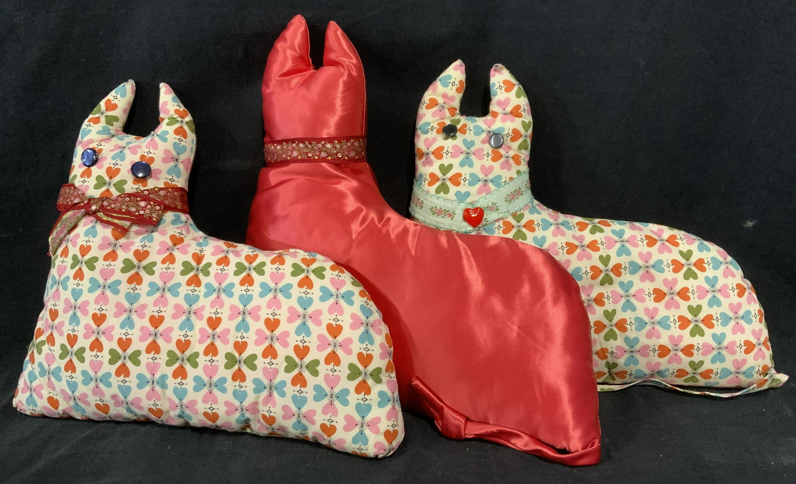 Lot 3 Upholstered Zoomorphic Cushions: Lot three vintage cushions, resemble animals of some kind. Two depict flower patterns. All three have ribbons wrapped around their necks. Two have eyes. Largest measures approx 11 inches x 11 inches. 