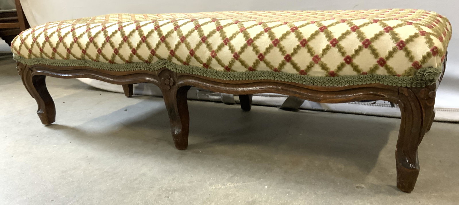 Vntg Upholstered Footstool W Scalloped Upholstery: Footstool is vintage possibly antique. Piece has cream, red & green toned upholstery. Piece has green toned scalloped edges. Base is wooden with carved detail. Piece measures approx 32 inches long 12 