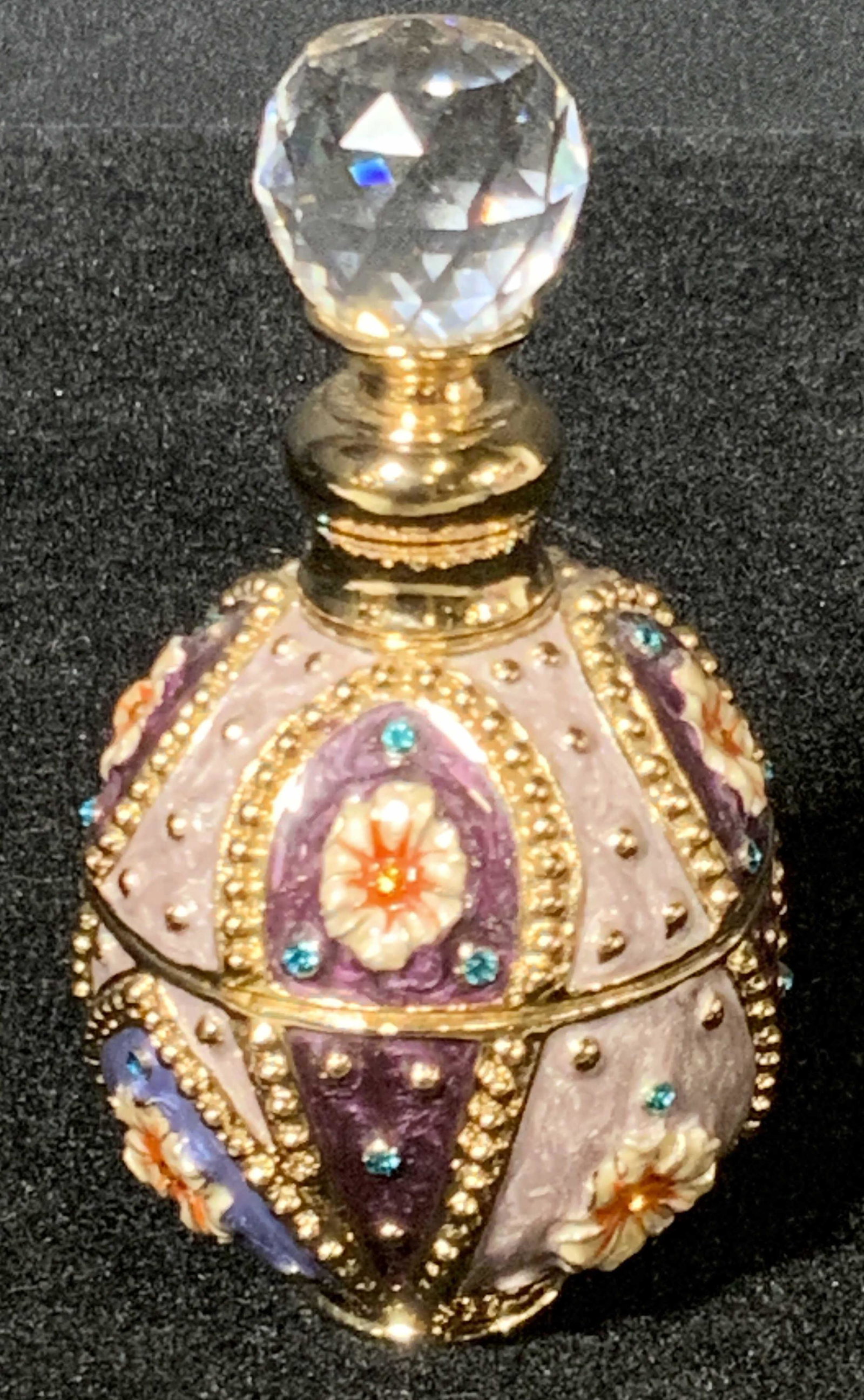 Floral Motif Bejeweled & Enamel Perfume Bottle: A metal perfume bottle that has been enameled and bejeweled. Circular in shape with floral motifs throughout the design. Stopper features a large faceted crystal. Piece has some weight to it. Jay Stro