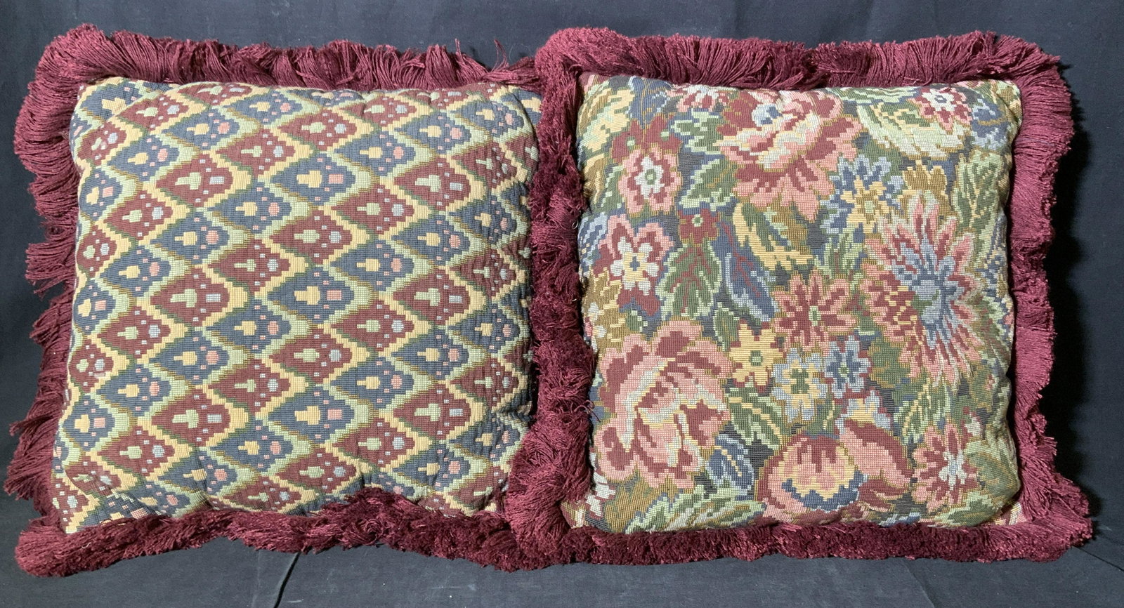 Pair Throw Pillows: Pair of fringed throw pillows. One decorated with a multi toned floral pattern, the other with a reticulated southwestern-like design. Fringes are wine toned, backs are royal blue velvet material. Eac