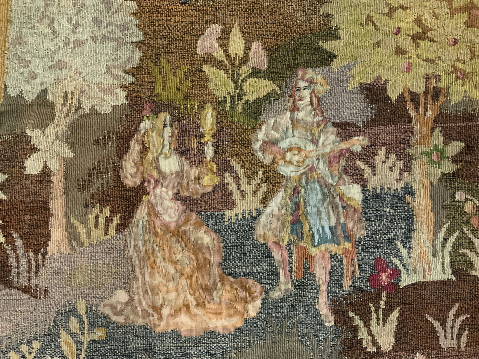 Antique Hand Loomed Tapestry: Antique hand loomed tapestry. Affixed to a metal Rod, ends or rod resemble fleur de lis, top and bottom of tapestry are fringed. Depicts two couples in, what appears to be, a garden. Wear is consisten