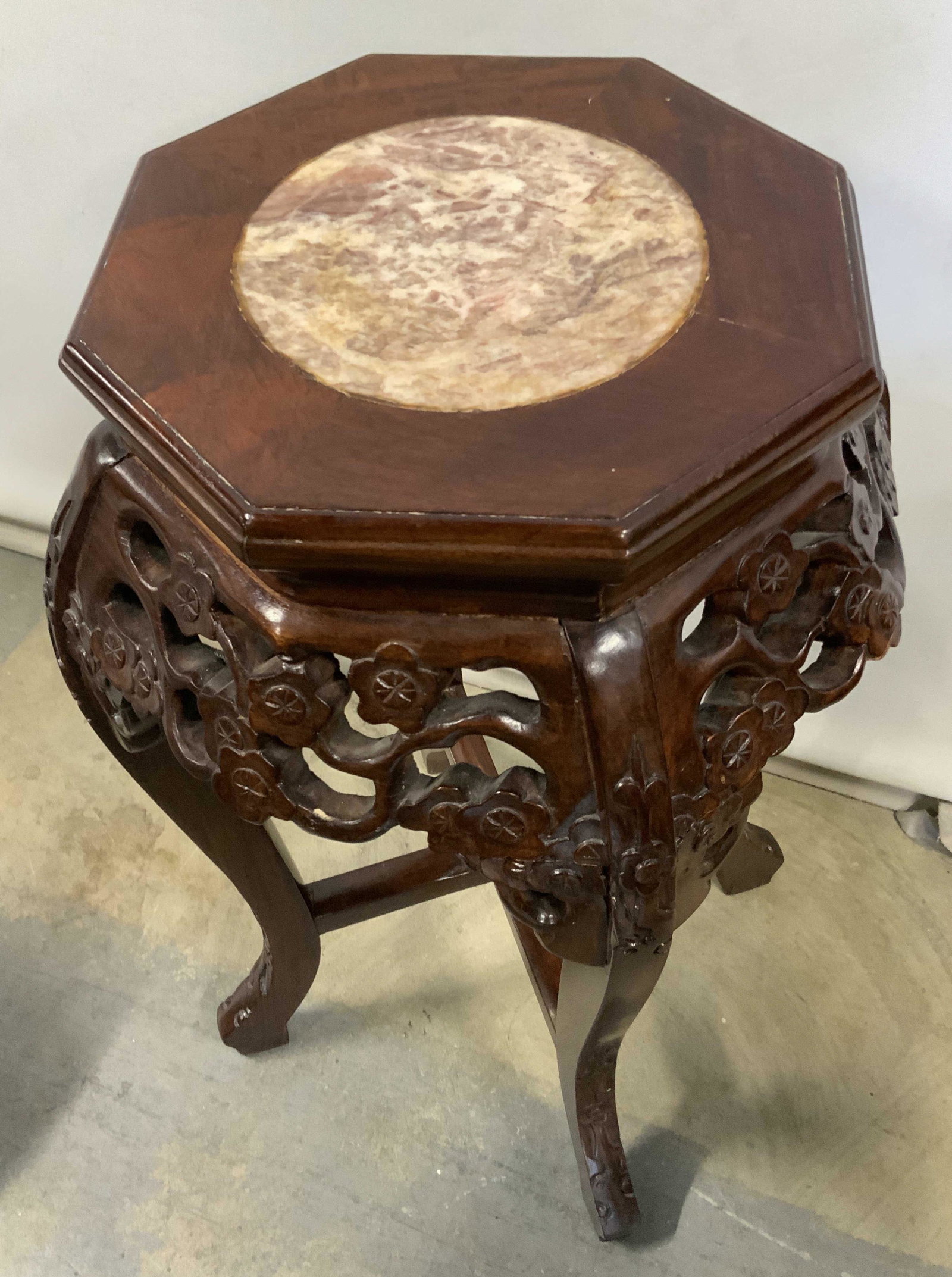 Vintage Marble Top Carved Wooden Plant Stand: A vintage plant stand crafted from marble and wood. Marble top center features earth toned hues. Woodwork is a rich brown hue with carved floral accents. Four legs with X shaped stretcher. Underside p